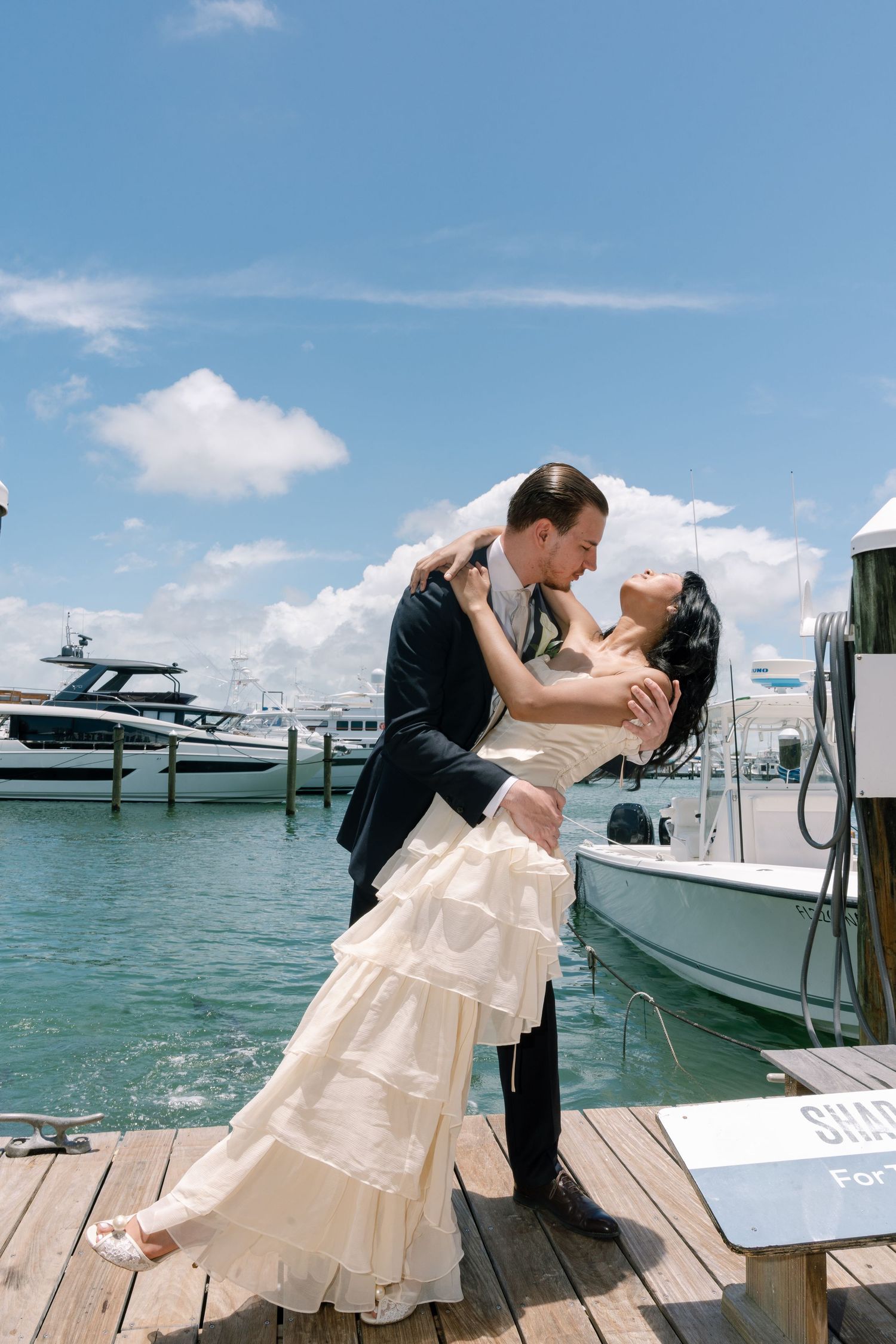 Bride and groom share a romantic dip kiss on a sunny marina dock with yachts and blue sky in the background.