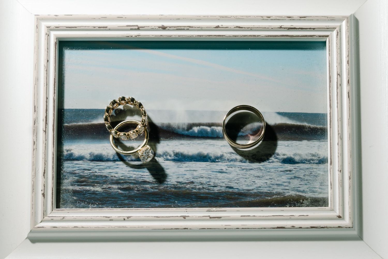 Framed photo of wedding rings displayed against ocean waves backdrop in white distressed frame.