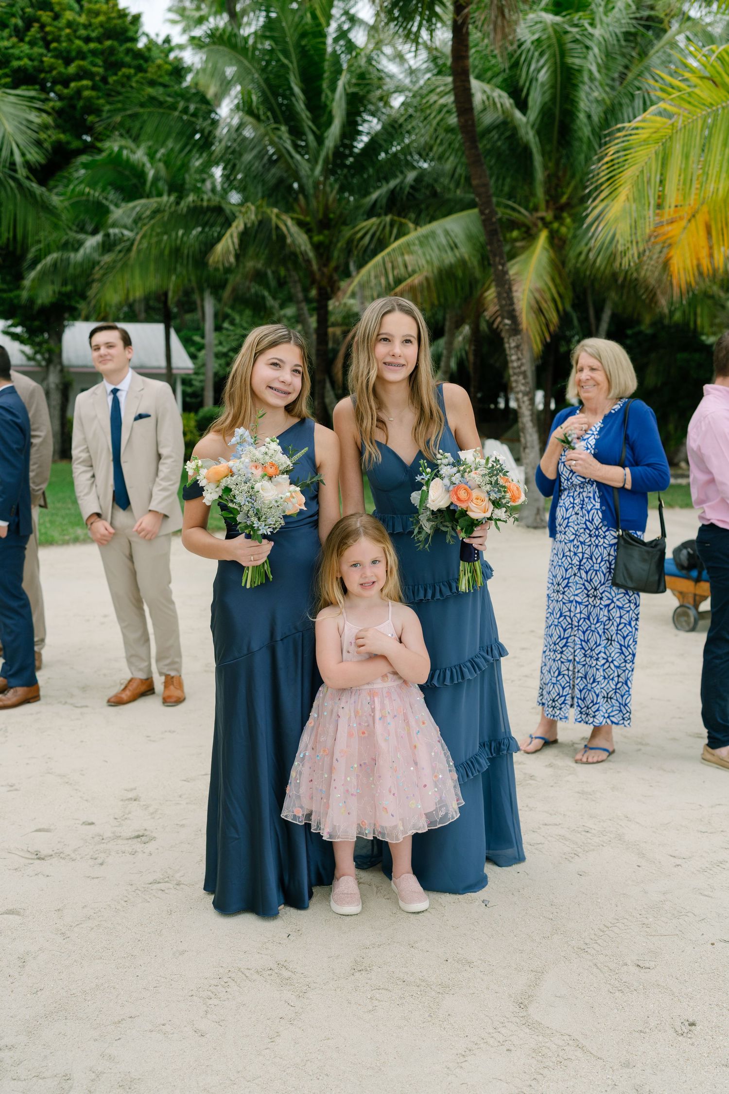 Wedding party members in navy blue dresses and formal attire gather on a tropical beach with palm trees.