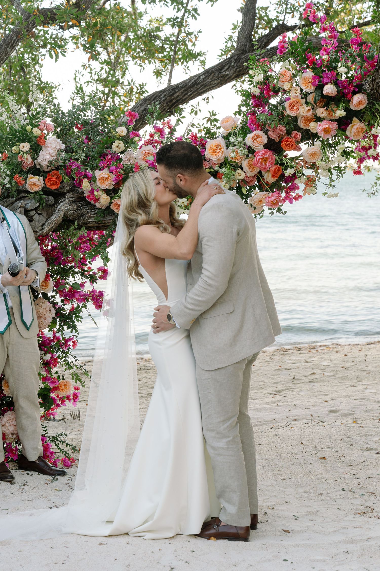 Beach wedding ceremony under a tree arch decorated with vibrant pink and coral flowers.