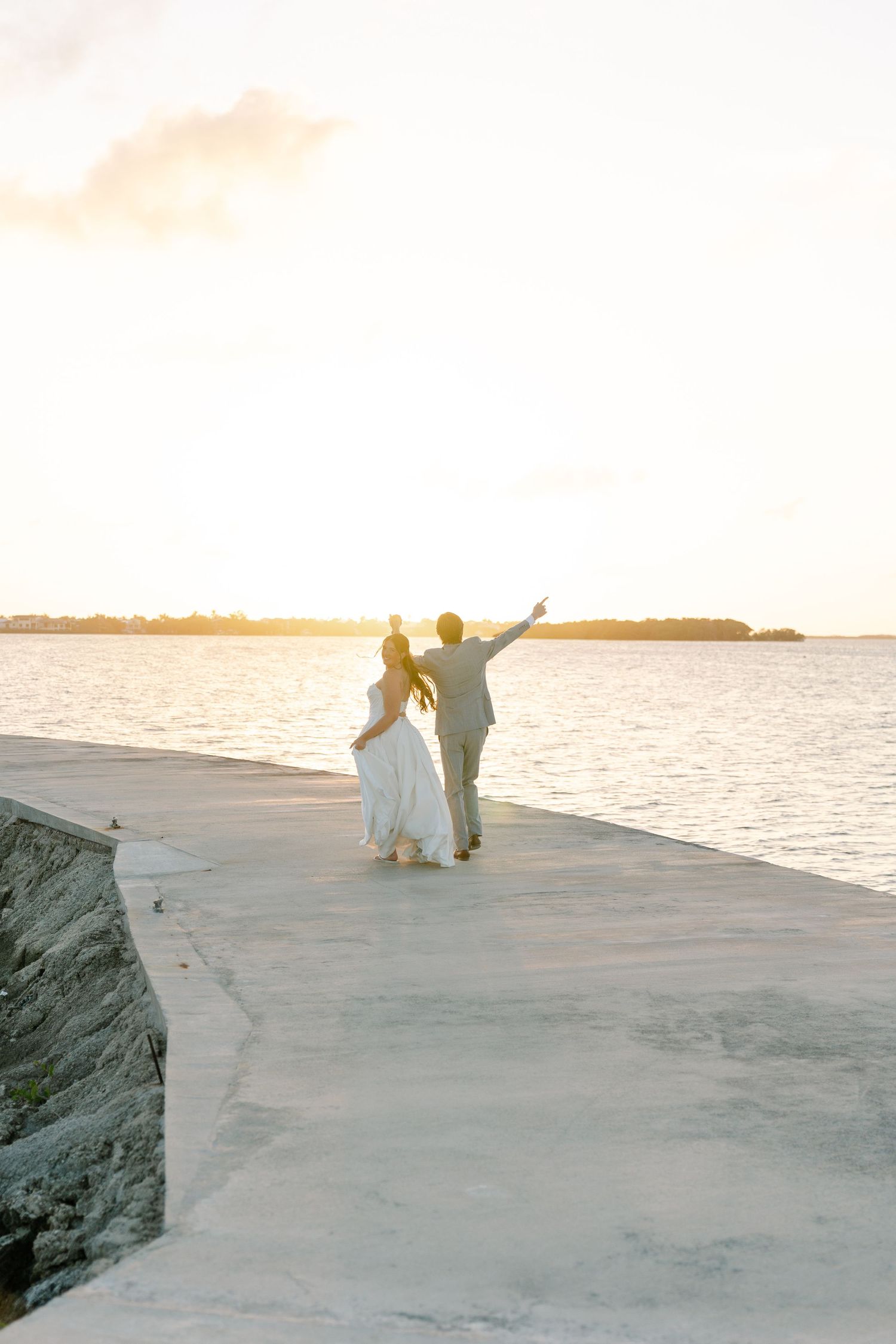 A couple walks hand-in-hand on a dock overlooking a body of water at sunset.