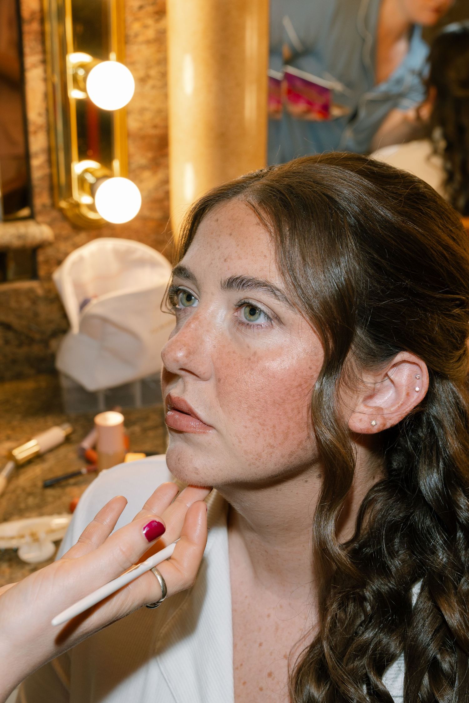 A woman with long curly hair applies makeup in front of a mirror with lights around it.