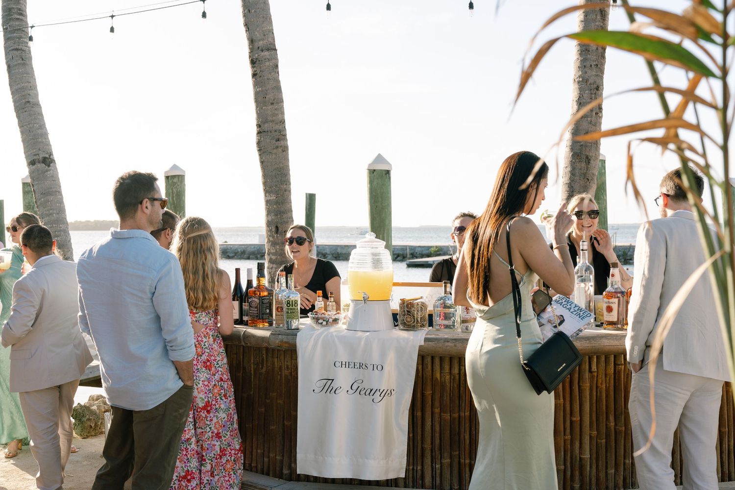 A group of people are gathered around a bar set up outdoors, with palm trees and buildings visible in the background.