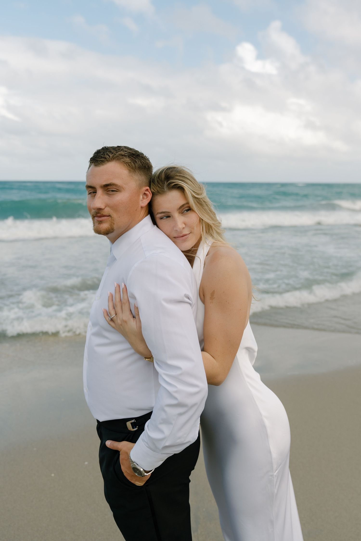 Couple in white wedding attire embraces romantically on sandy beach with ocean waves behind them.