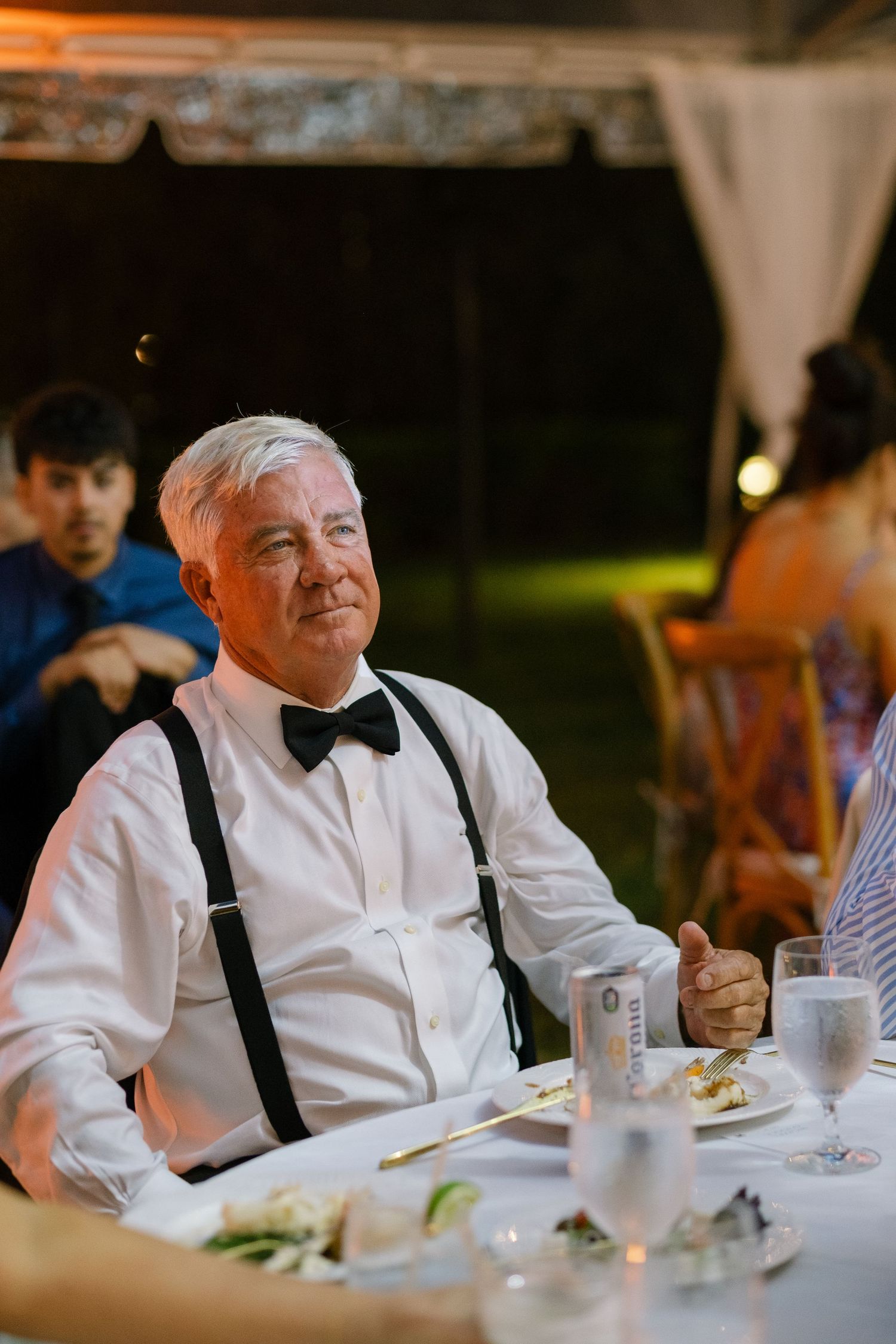 Elderly man in white shirt with suspenders and bow tie smiling at a wedding reception dinner table.