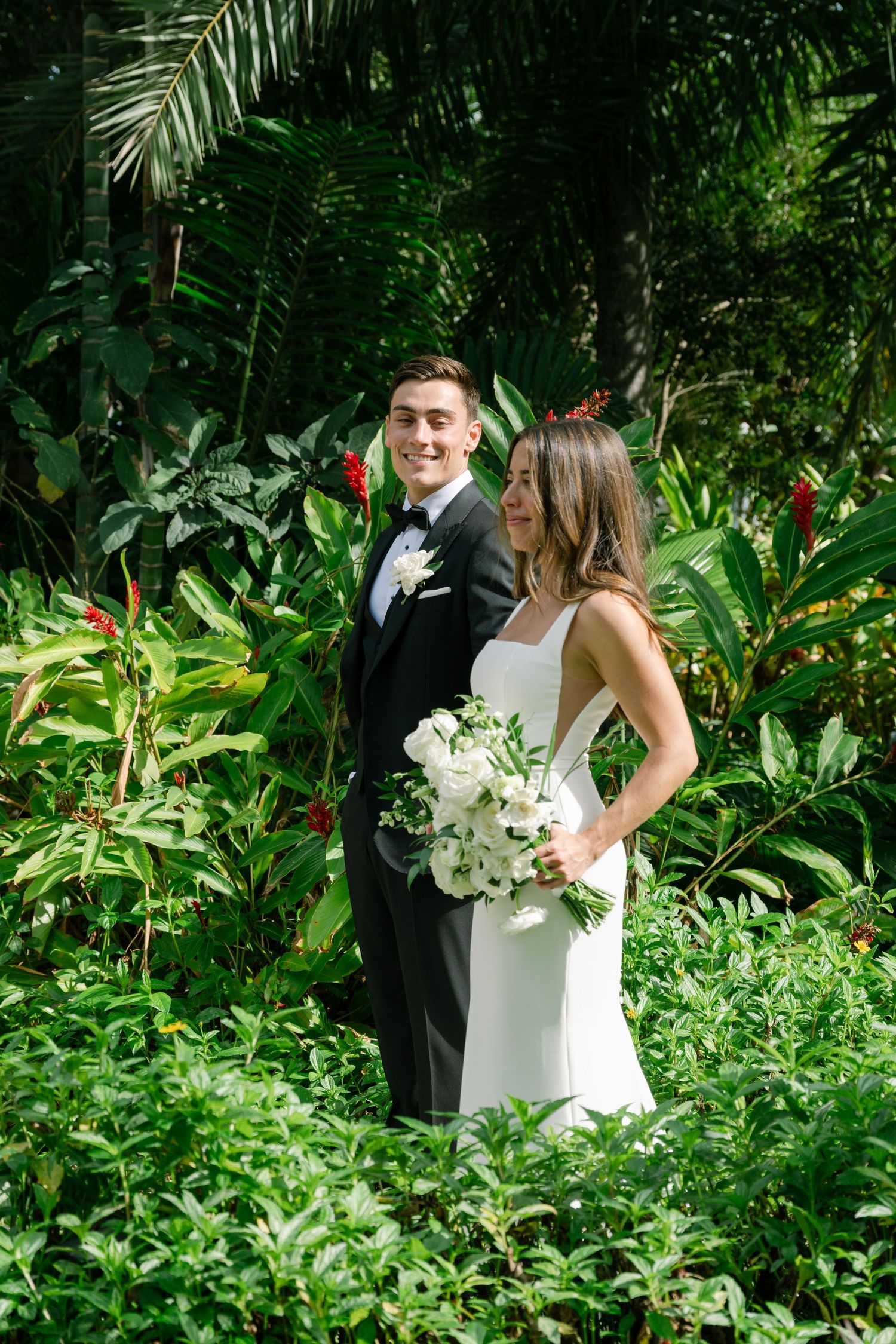 Bride and groom posing together in a lush tropical garden, bride holding white bouquet.
