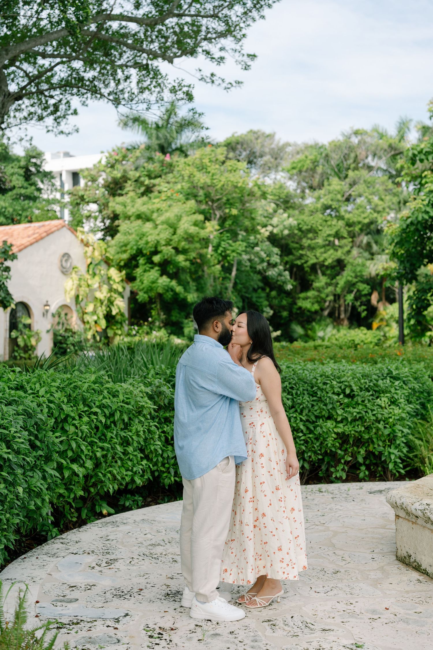 Couple sharing a romantic kiss in a lush garden setting, man in blue shirt and woman in floral dress.