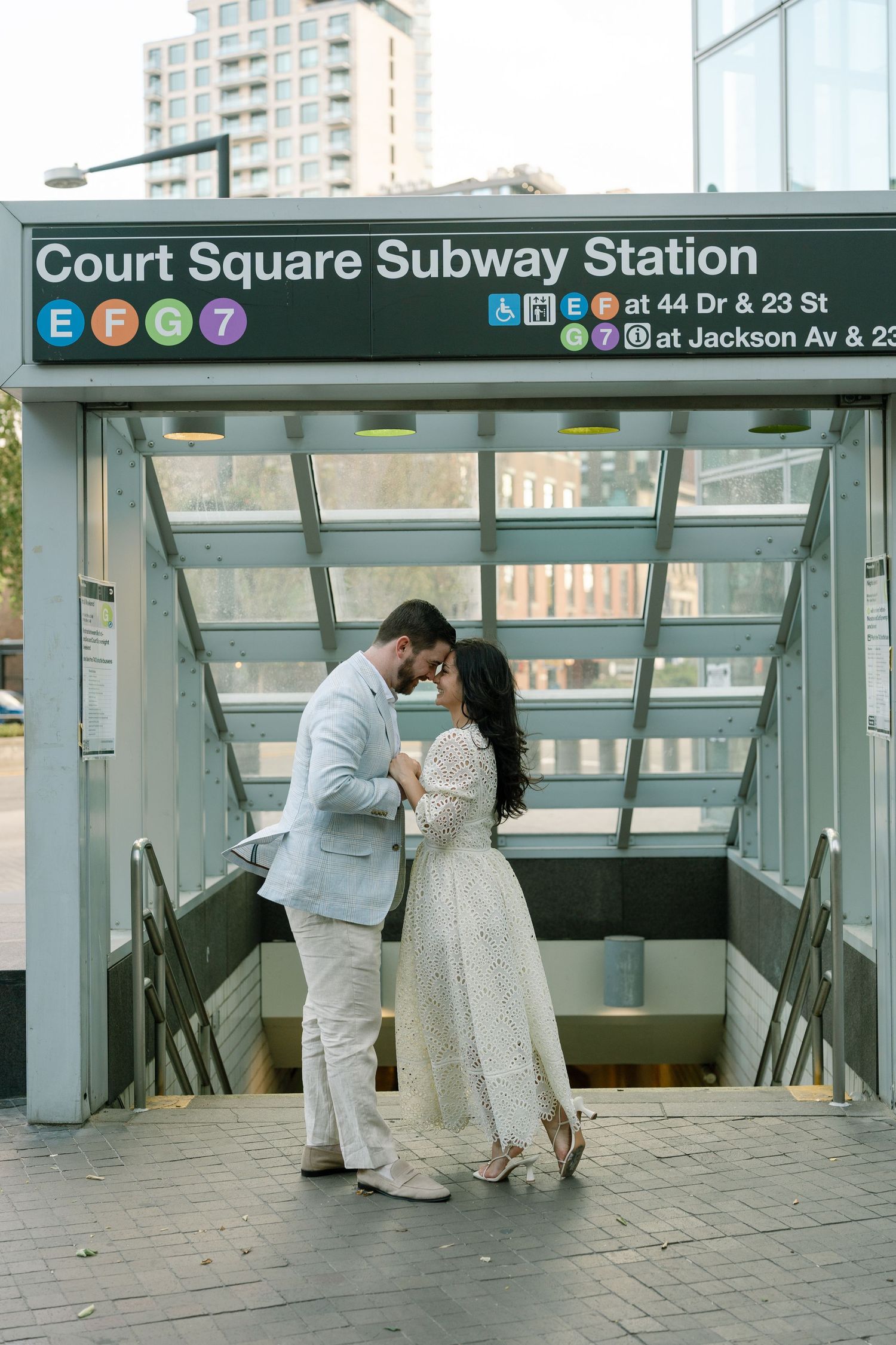 Couple sharing romantic moment at Court Square Subway Station entrance in NYC, dressed in elegant white attire.