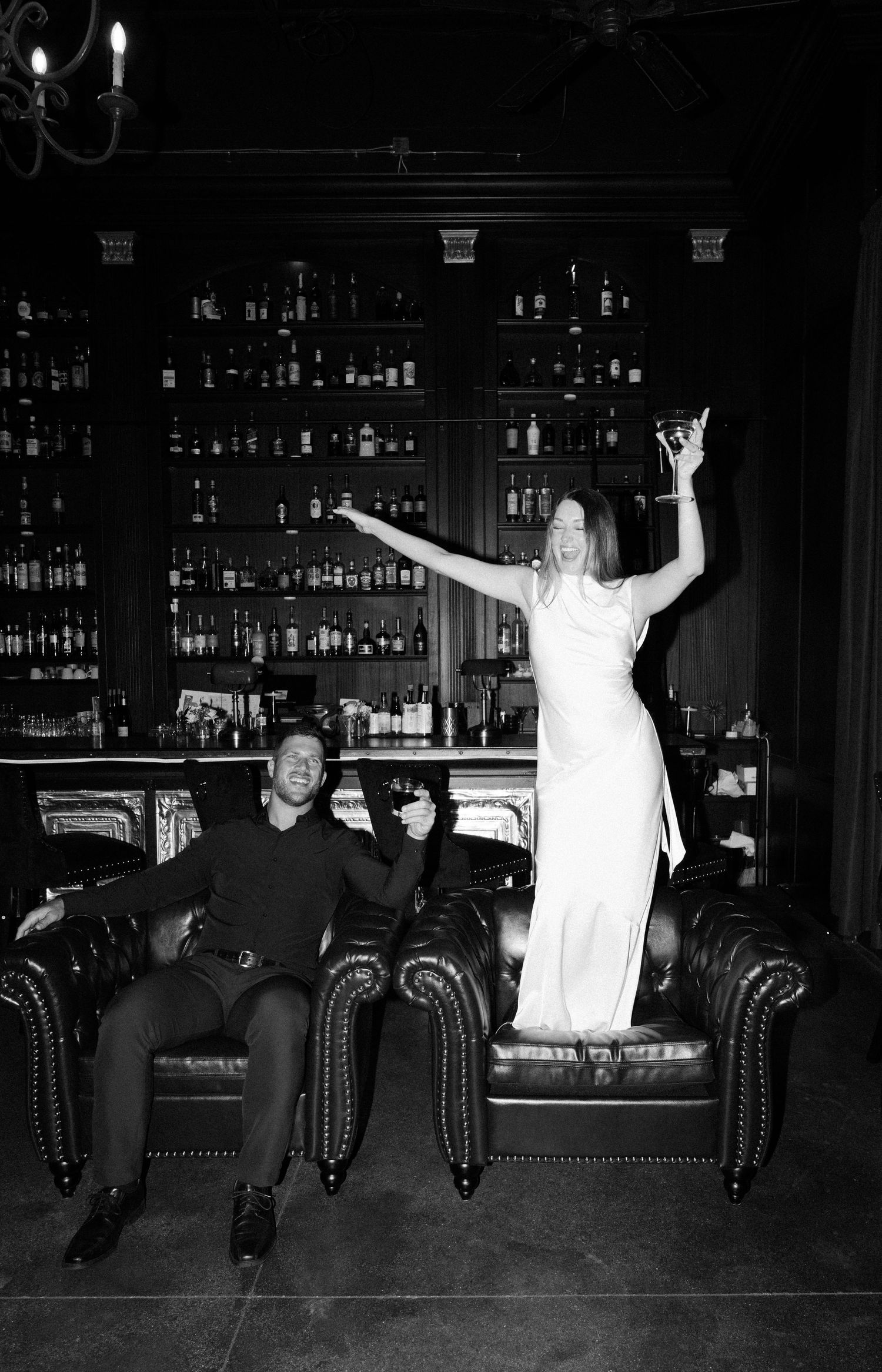 Four black and white photos show people sitting and jumping on a leather couch at a bar with bottles behind them.