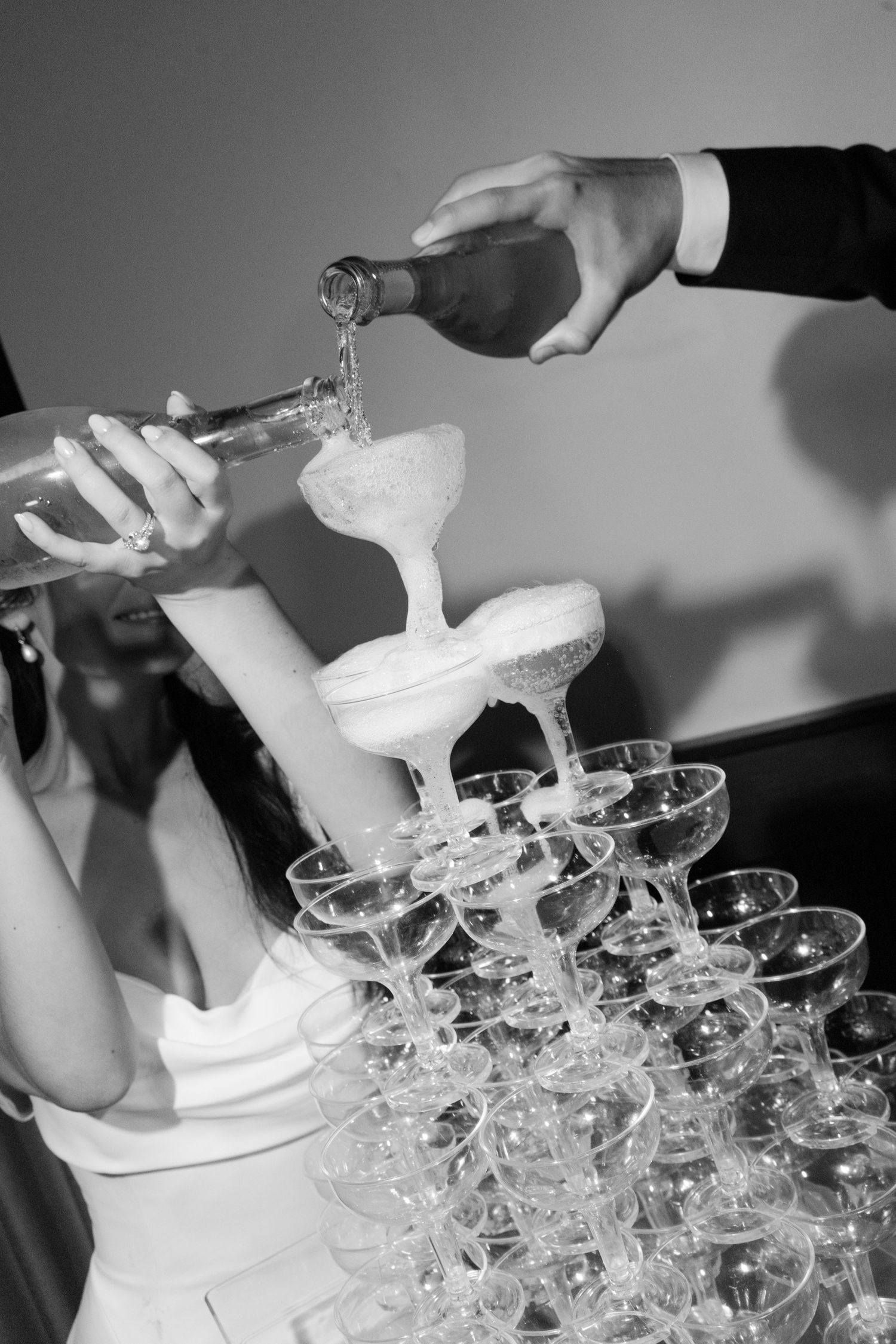 Black and white photo of champagne tower being filled at wedding reception with two people pouring bottles.