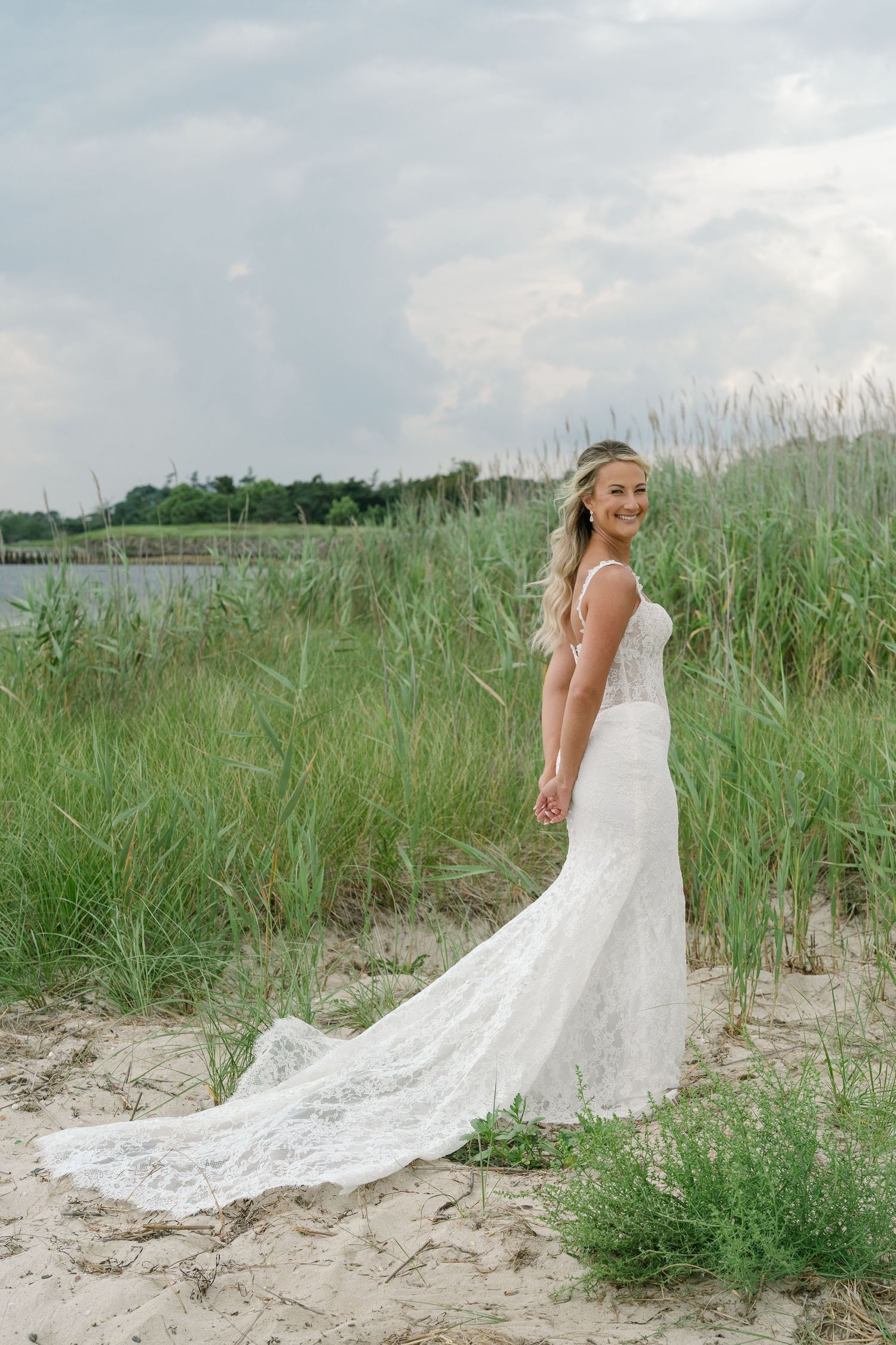 Radiant bride in a lace mermaid wedding gown with a long train stands on a sandy beach among tall grasses.