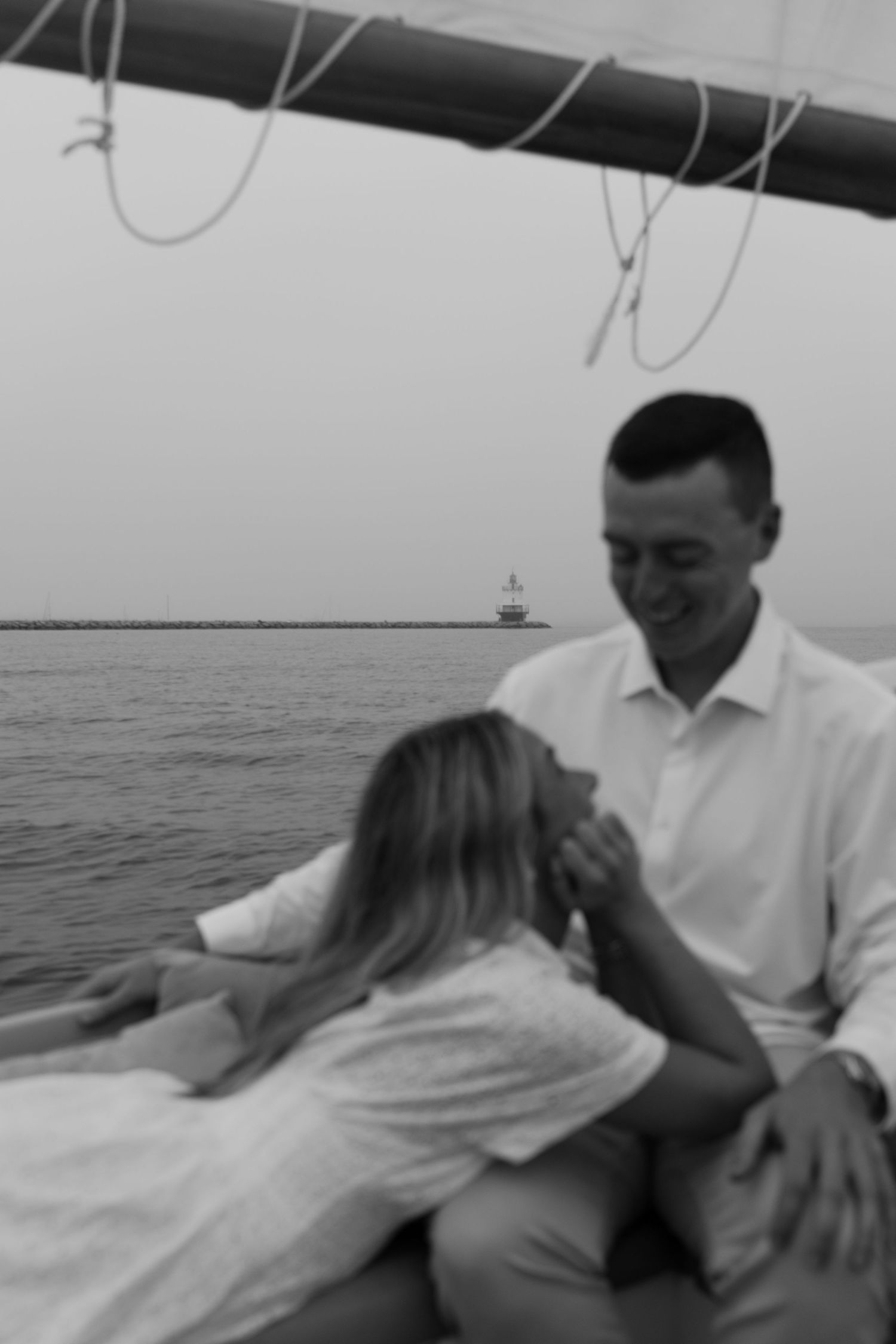 Black and white photo of a couple sharing a romantic moment on a sailboat with a lighthouse in the background.