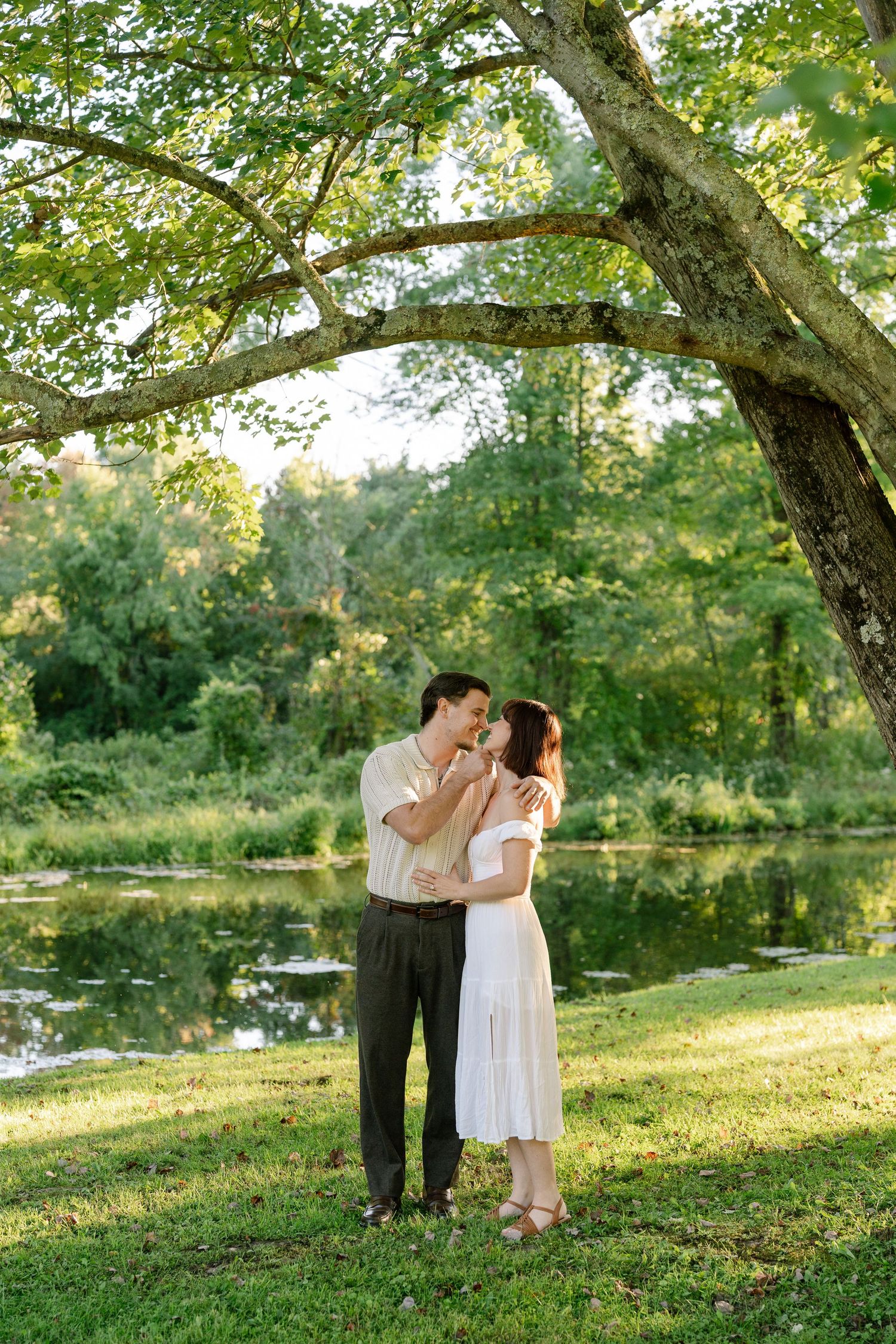 Couple sharing a romantic moment under a large tree by a serene pond in a lush green park setting.