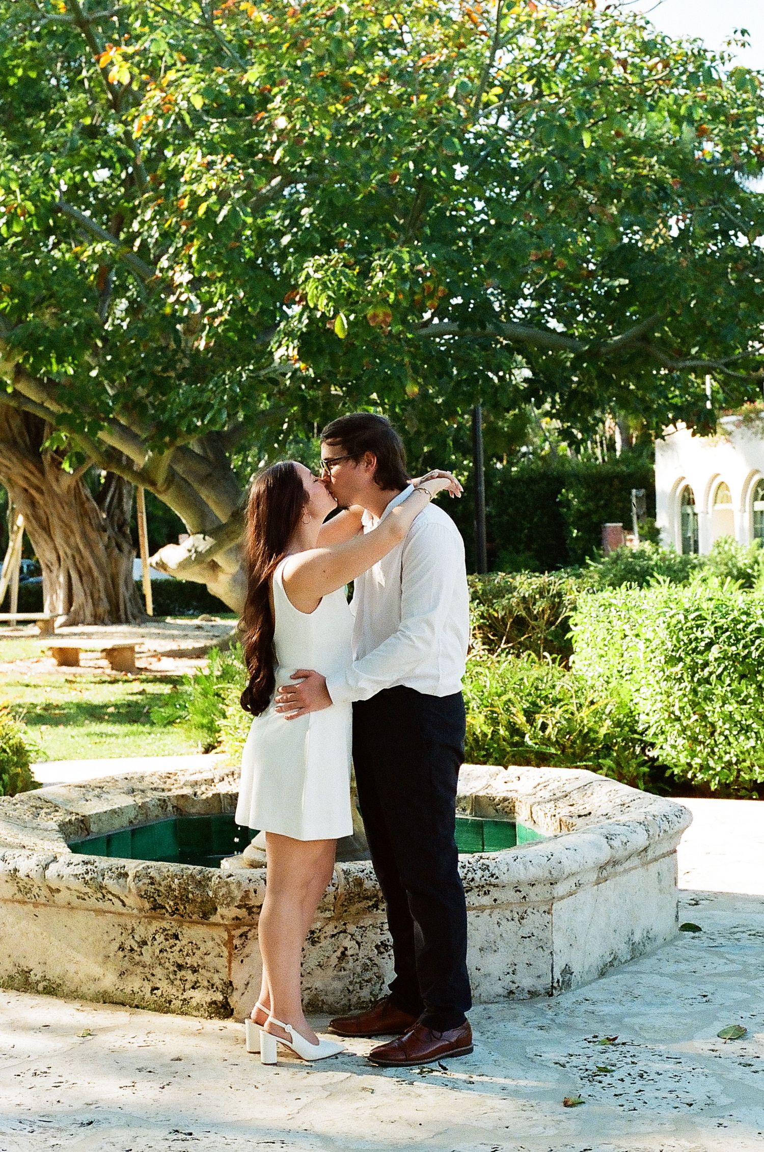 A romantic couple shares a tender moment on a stone wall in a lush garden setting during a sunny day.