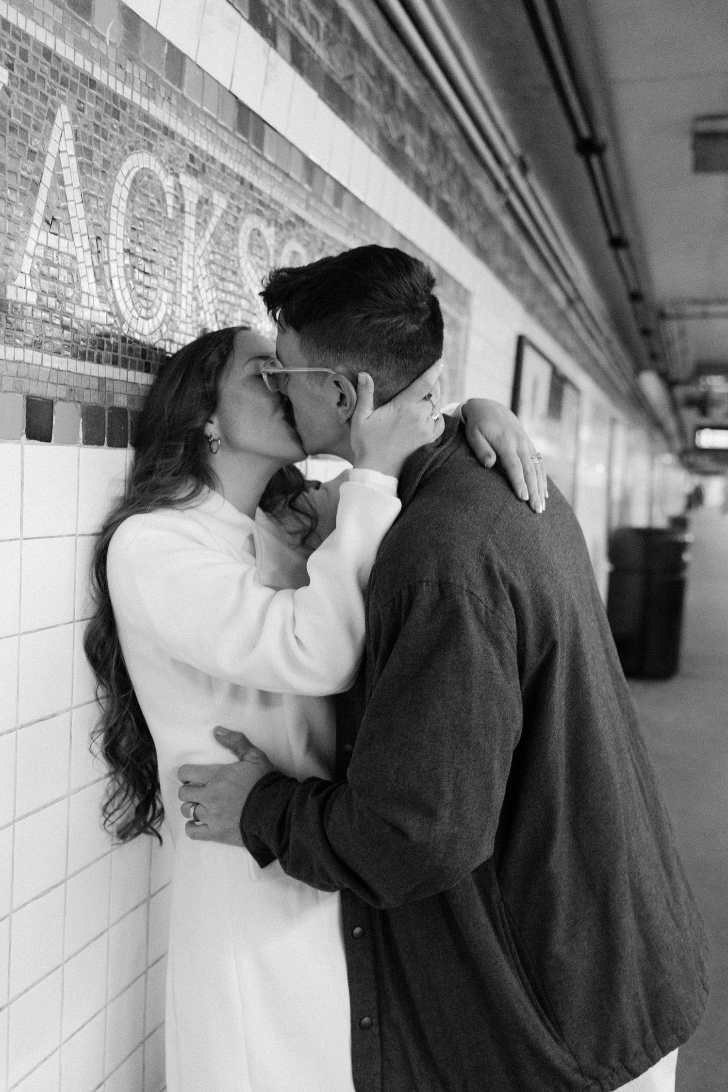 A couple shares an intimate moment against a white tiled wall in a dimly lit corridor.