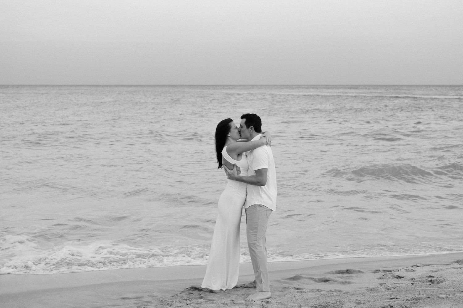 A couple embraces on a beach in black and white as waves roll in behind them.