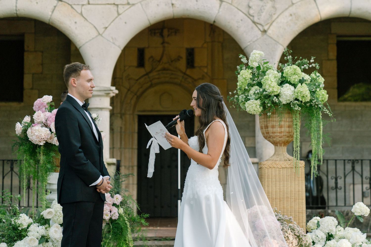 Bride in white gown reads vows to groom before stone arched chapel with lush floral arrangements.
