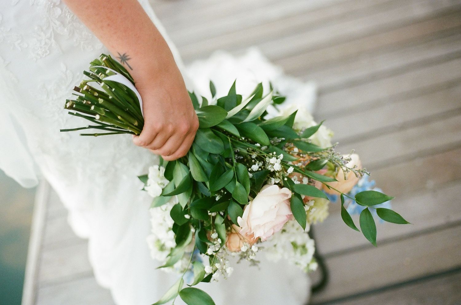 A hand arranges fresh white flowers and green philodendron leaves in a vase against a light gray background.