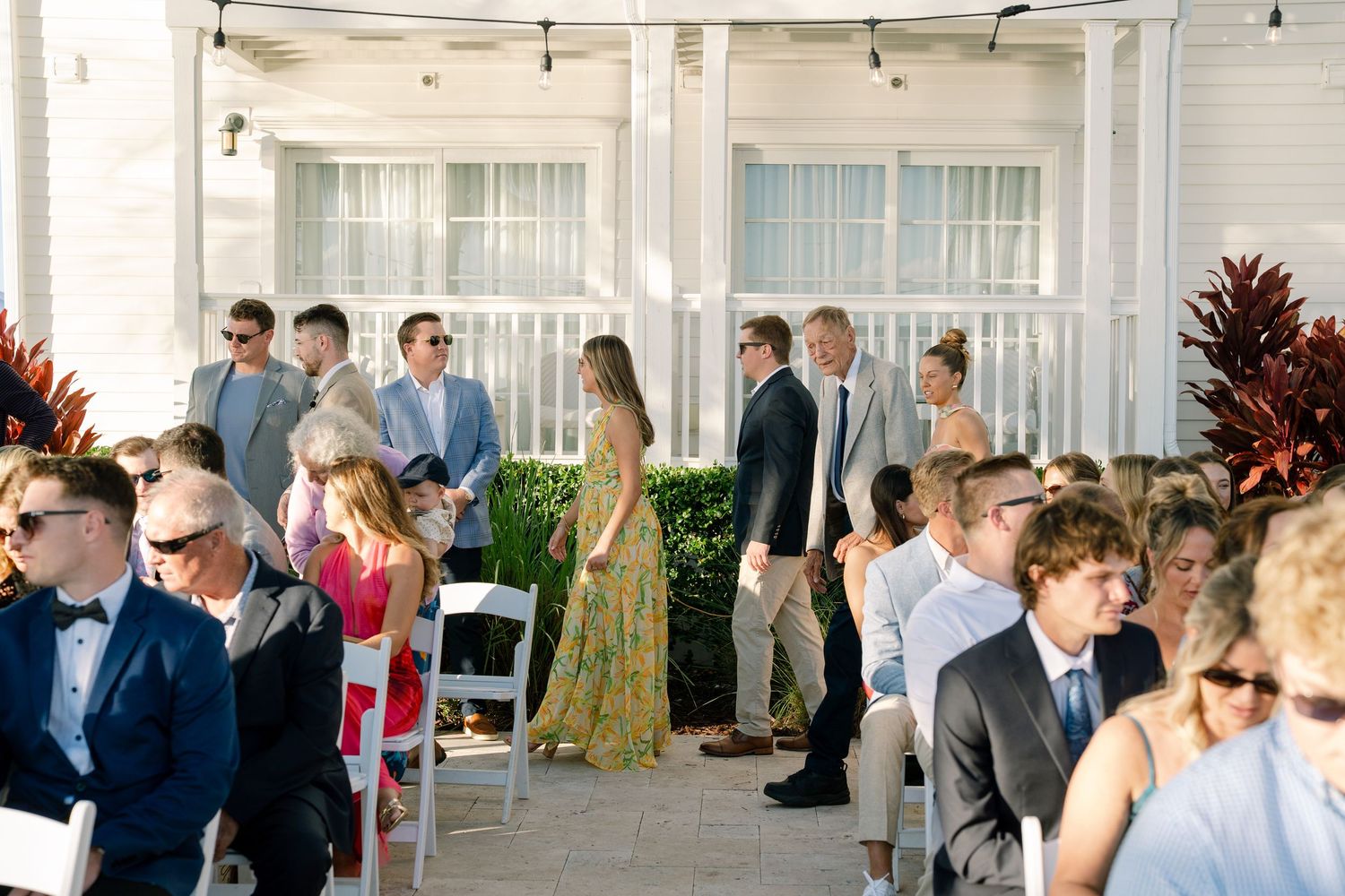 Wedding guests stand in rows outdoors during a daytime ceremony with string lights overhead and white building in background.