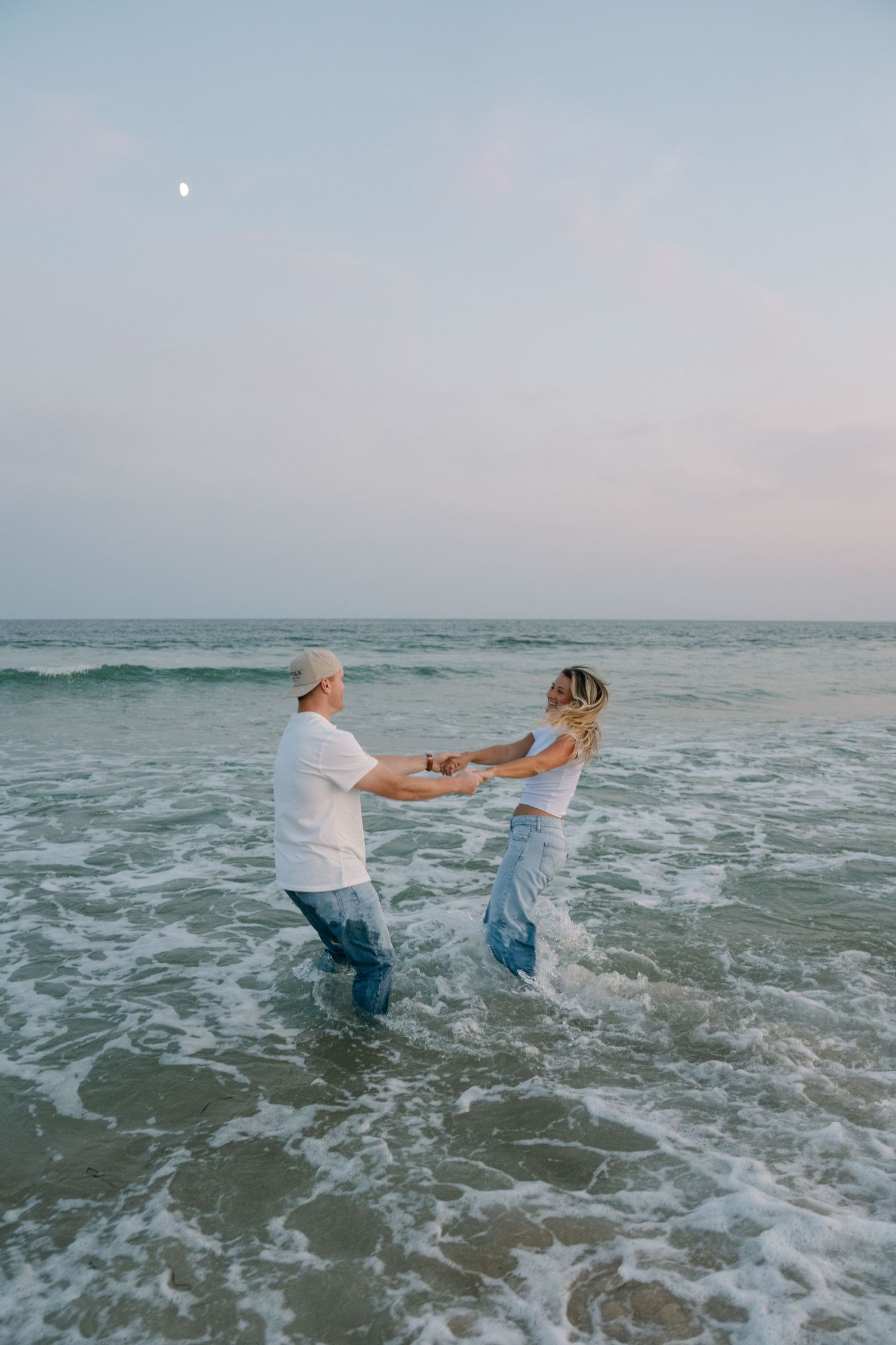 Two people hold hands while playing in the ocean waves at sunset, creating a joyful beach moment.