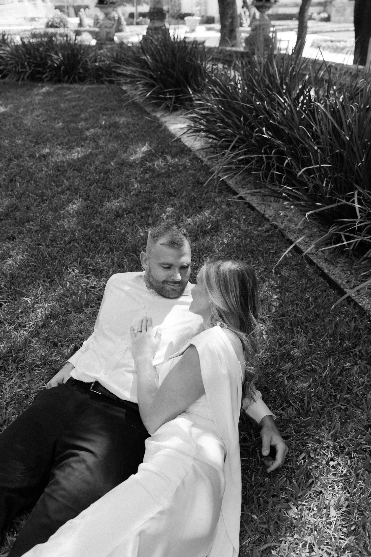 A couple in wedding attire lying together on grass in a romantic black and white photograph.