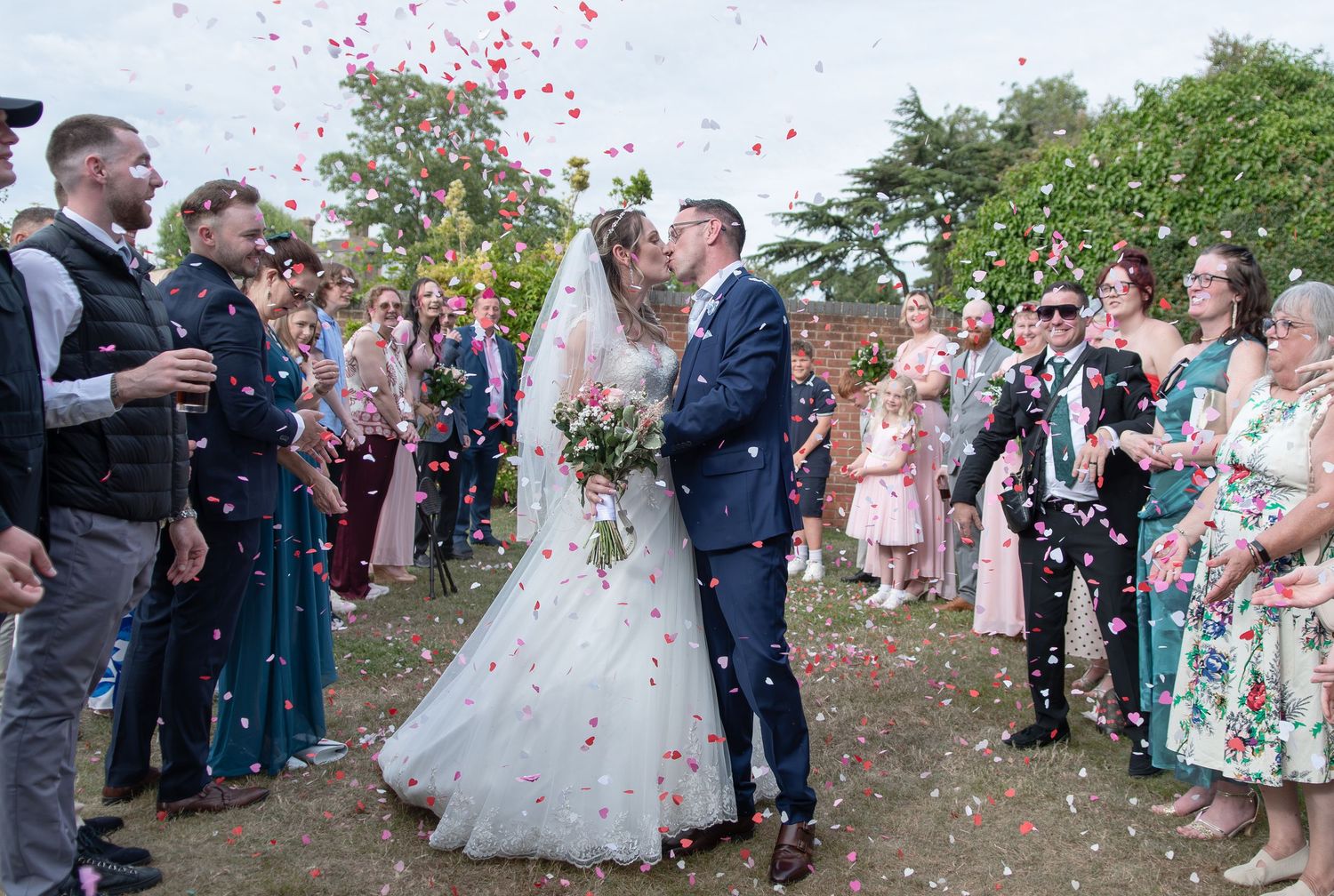Bride and groom kissing during outdoor wedding confetti shower surrounded by smiling guests in summer garden celebration.