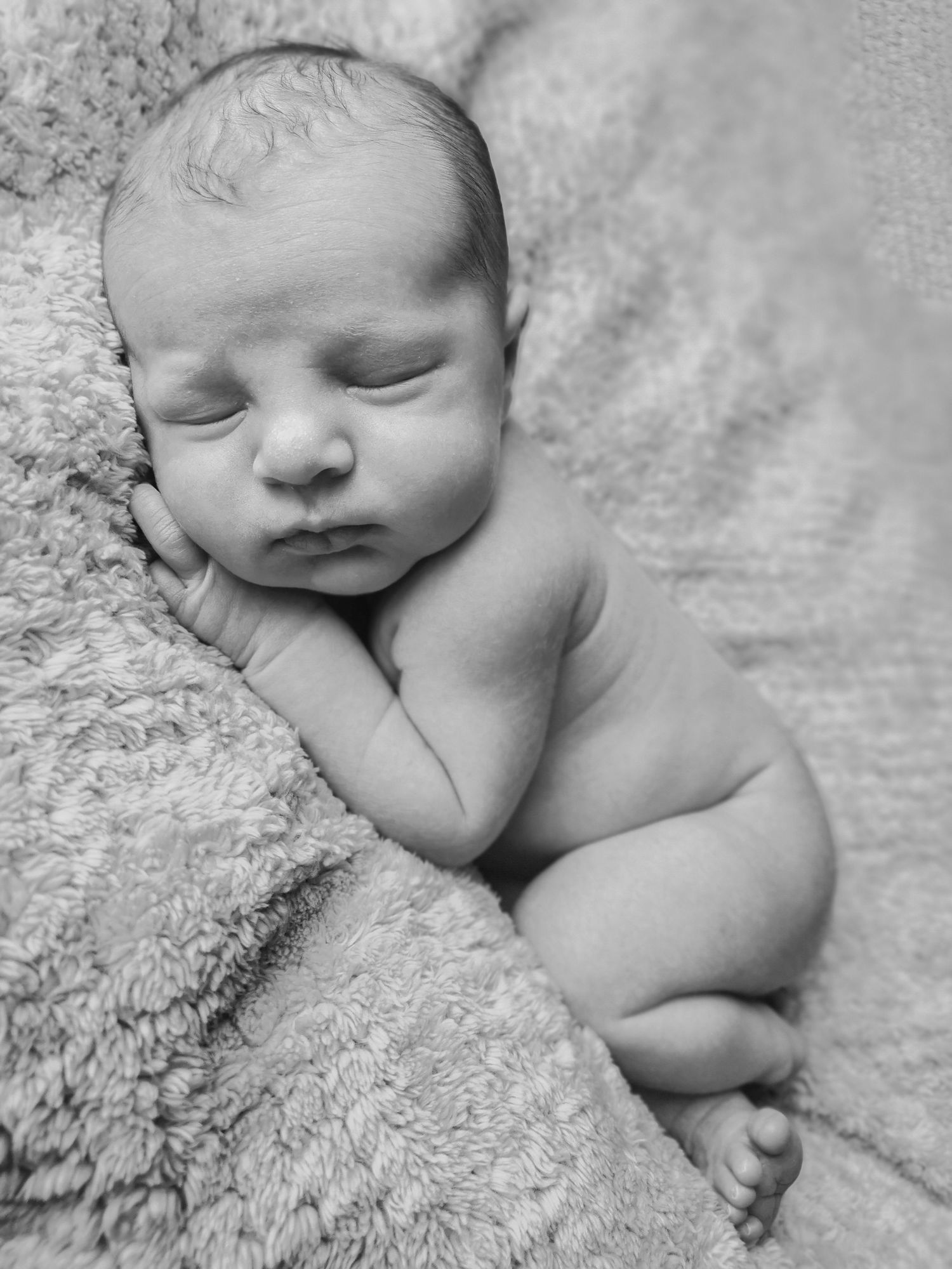 Black and white photo of naked newborn baby sleeping curled up on soft textured blanket.