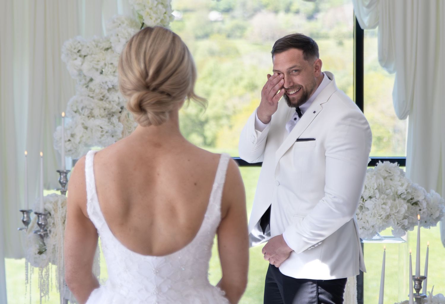 Emotional groom in white suit wiping tears as bride in backless lace wedding dress approaches altar.
