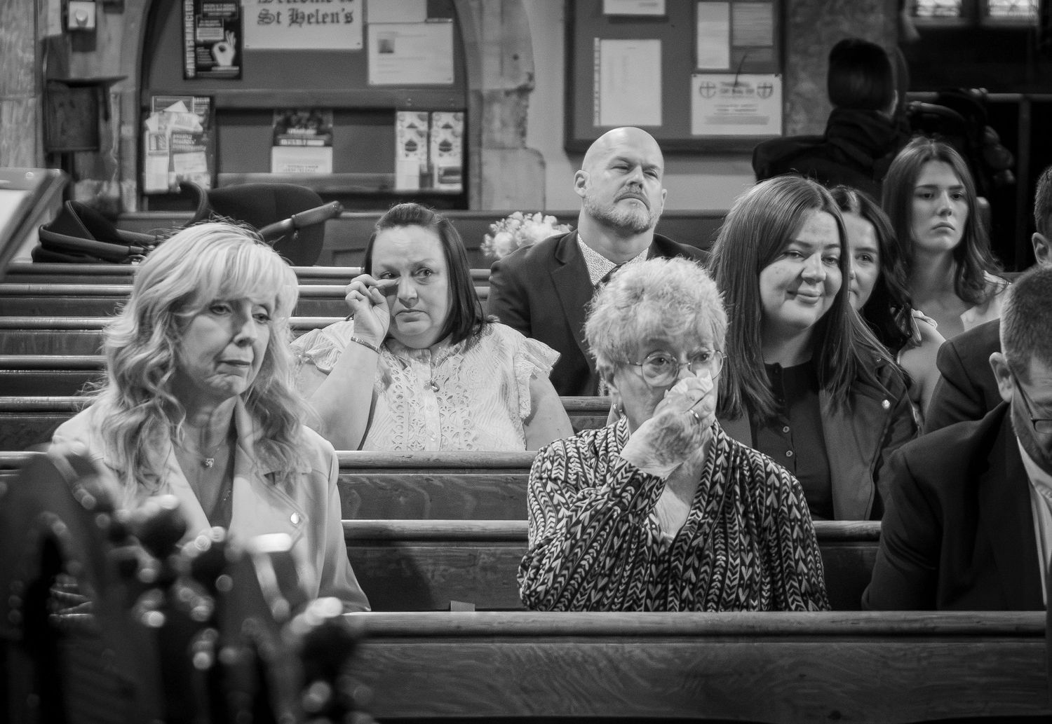 Black and white photo of congregation in church pews, elderly woman wiping tears during an emotional ceremony.