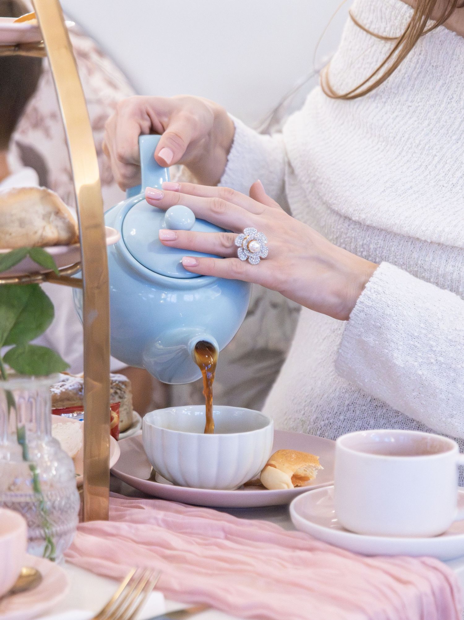 Woman in white sweater pouring tea from a blue teapot into a pink cup at an elegant tea party setting.