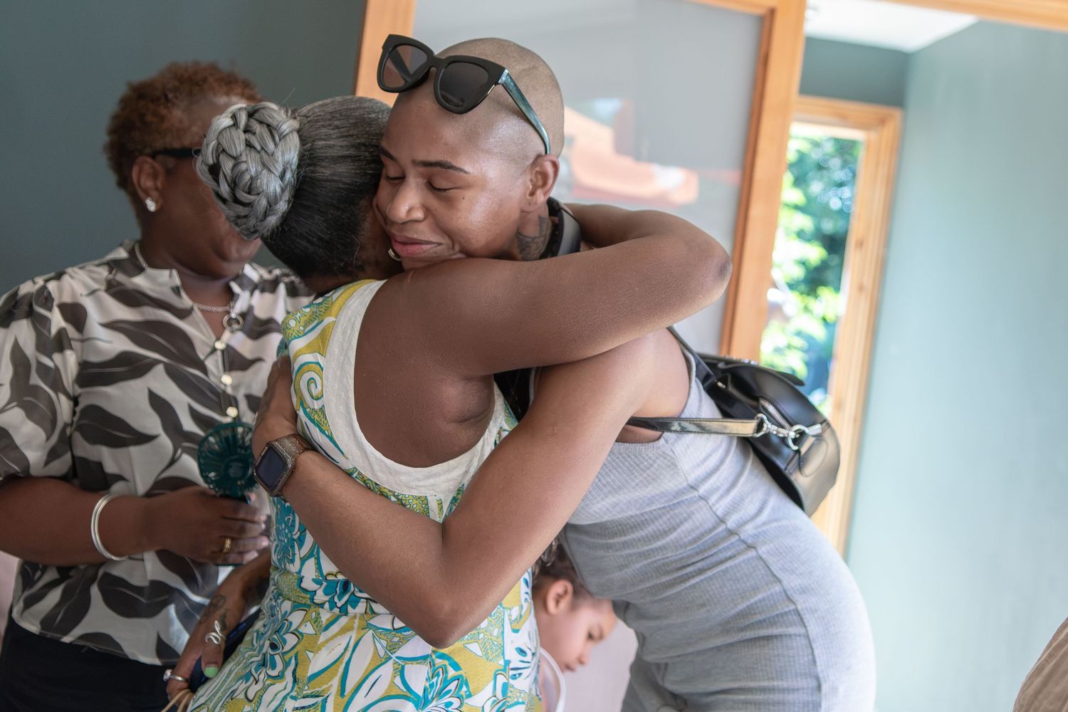 Two women share a warm embrace indoors while another woman stands nearby in the background.