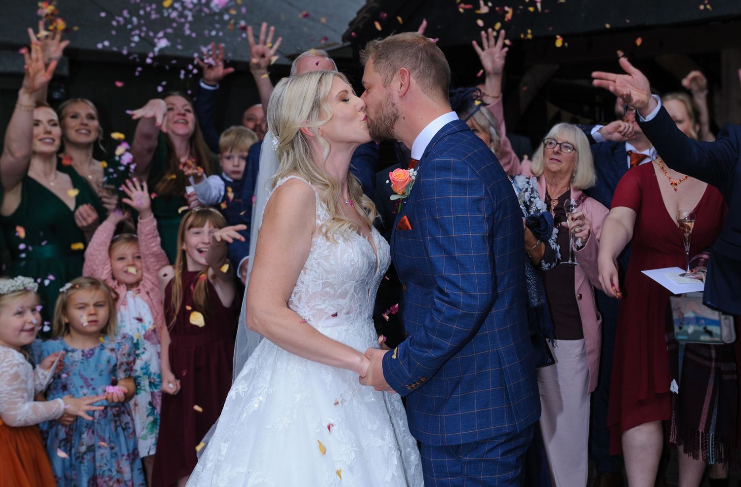 Bride and groom share a kiss surrounded by guests throwing colorful confetti at an outdoor wedding celebration.