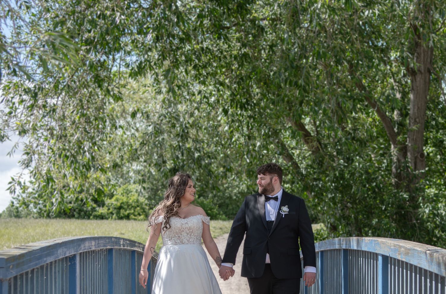 A bride and groom hold hands walking on a bridge surrounded by lush green trees on their wedding day.