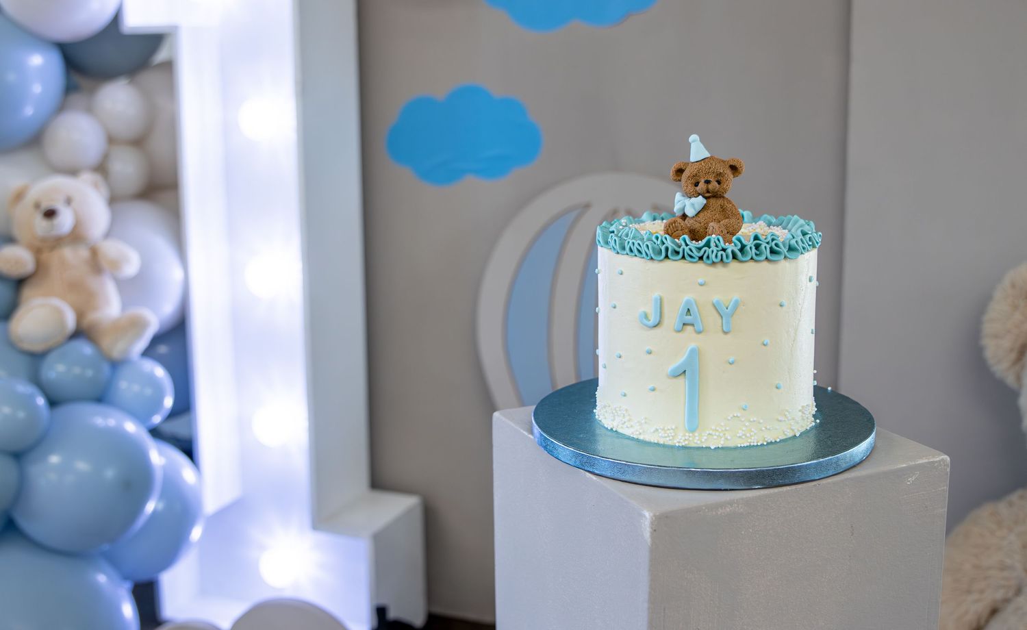 First birthday cake with blue theme featuring teddy bear topper and name Jay, displayed on white pedestal with balloon decorations.