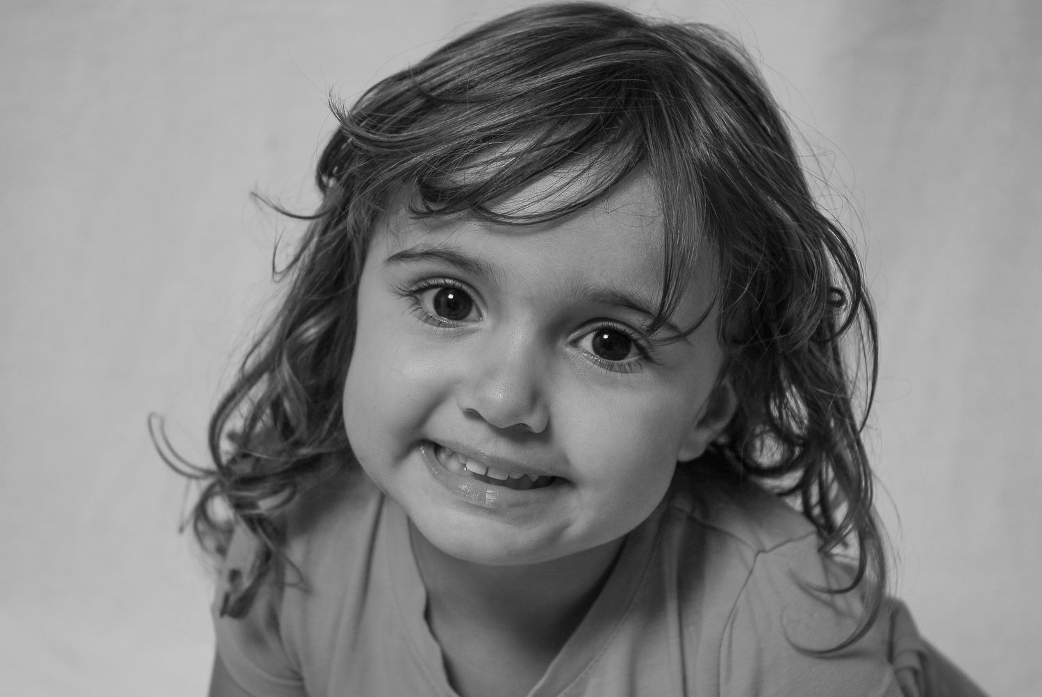Black and white portrait of a cute toddler girl with curly hair and big dark eyes smiling at the camera.