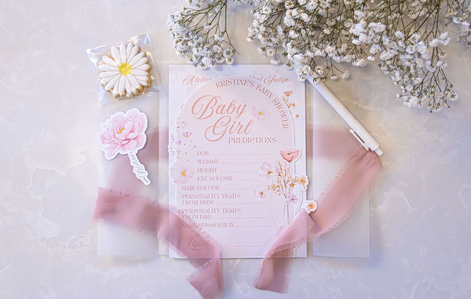 Pink baby girl shower invitation flatlay with floral decorations, ribbon, and pen on marble surface.