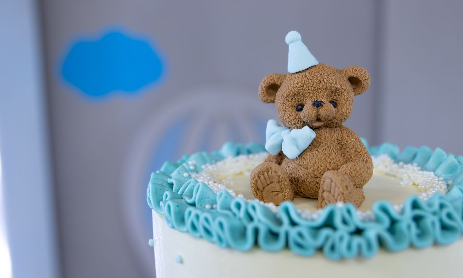 A cute fondant teddy bear wearing a blue party hat and bow tie sits atop a white birthday cake with blue ruffles.