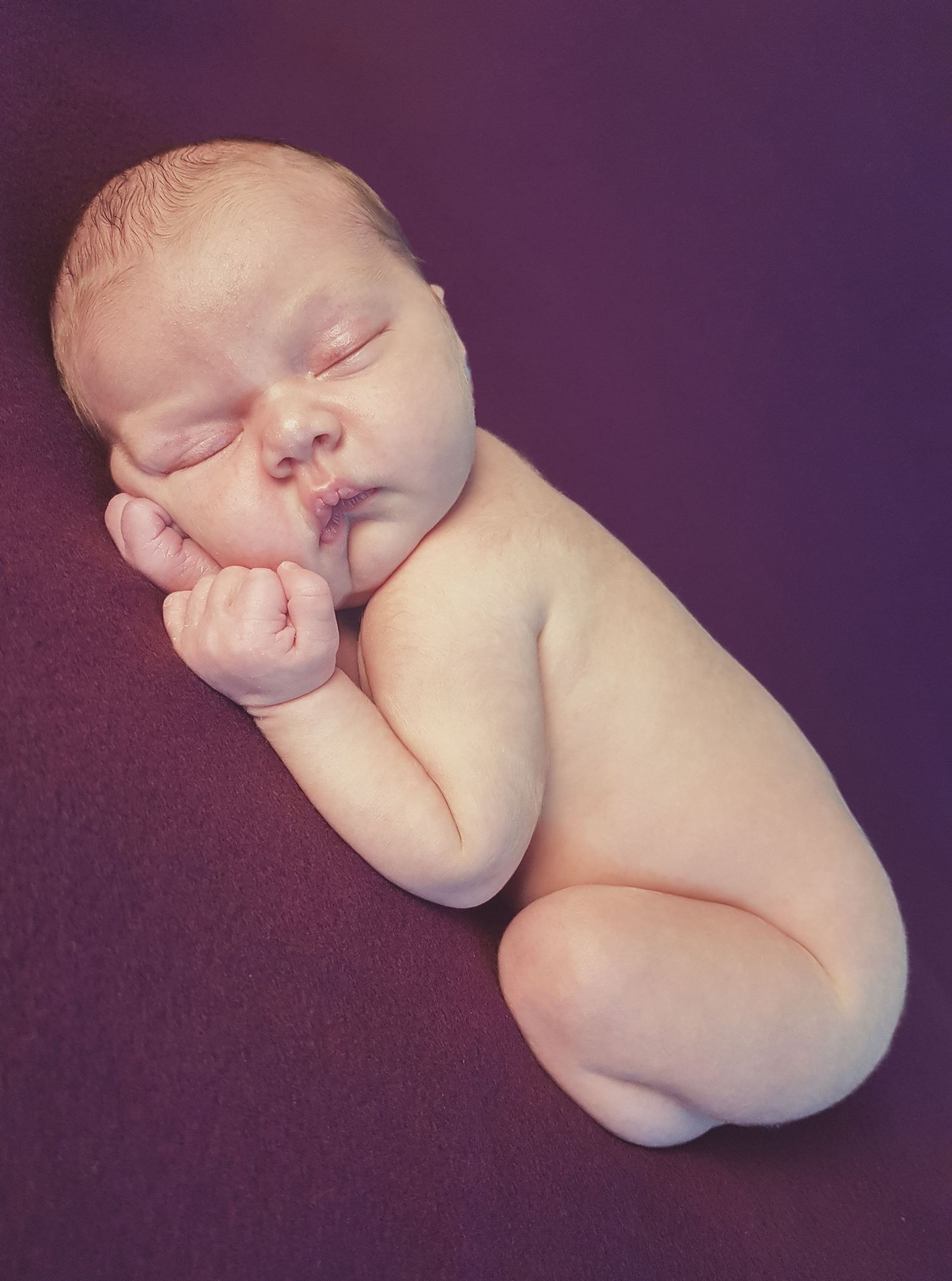Peaceful newborn baby sleeping curled up on a purple background in a classic newborn photography pose.