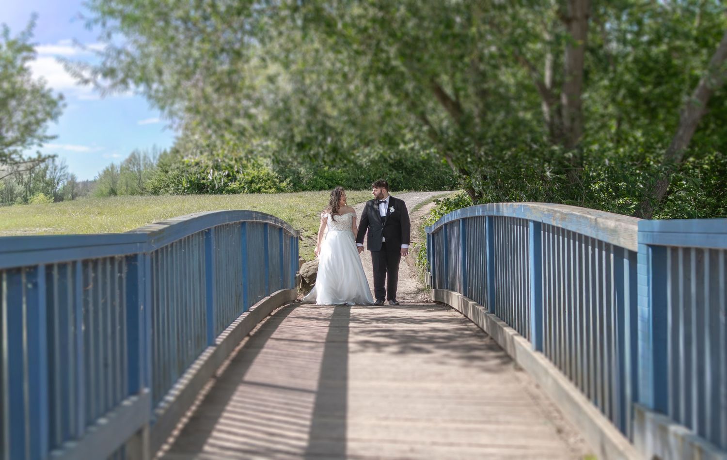 Bride and groom walking hand in hand on a blue wooden bridge surrounded by lush green trees on a sunny wedding day.