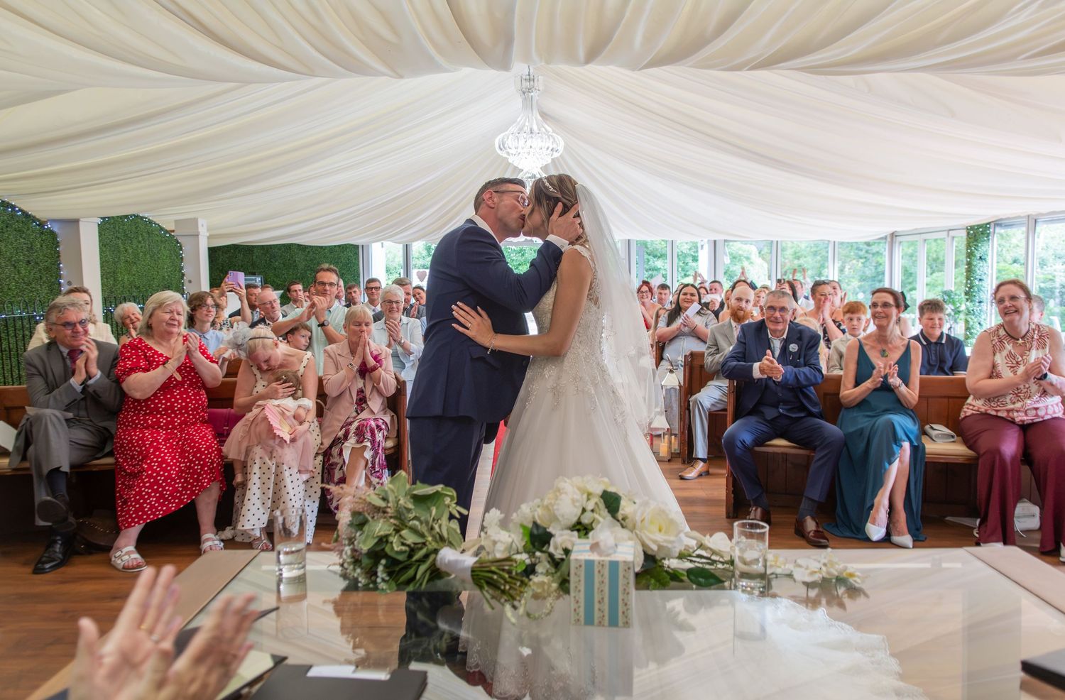 A bride and groom share their first kiss at the altar as wedding guests applaud in a elegant marquee venue.