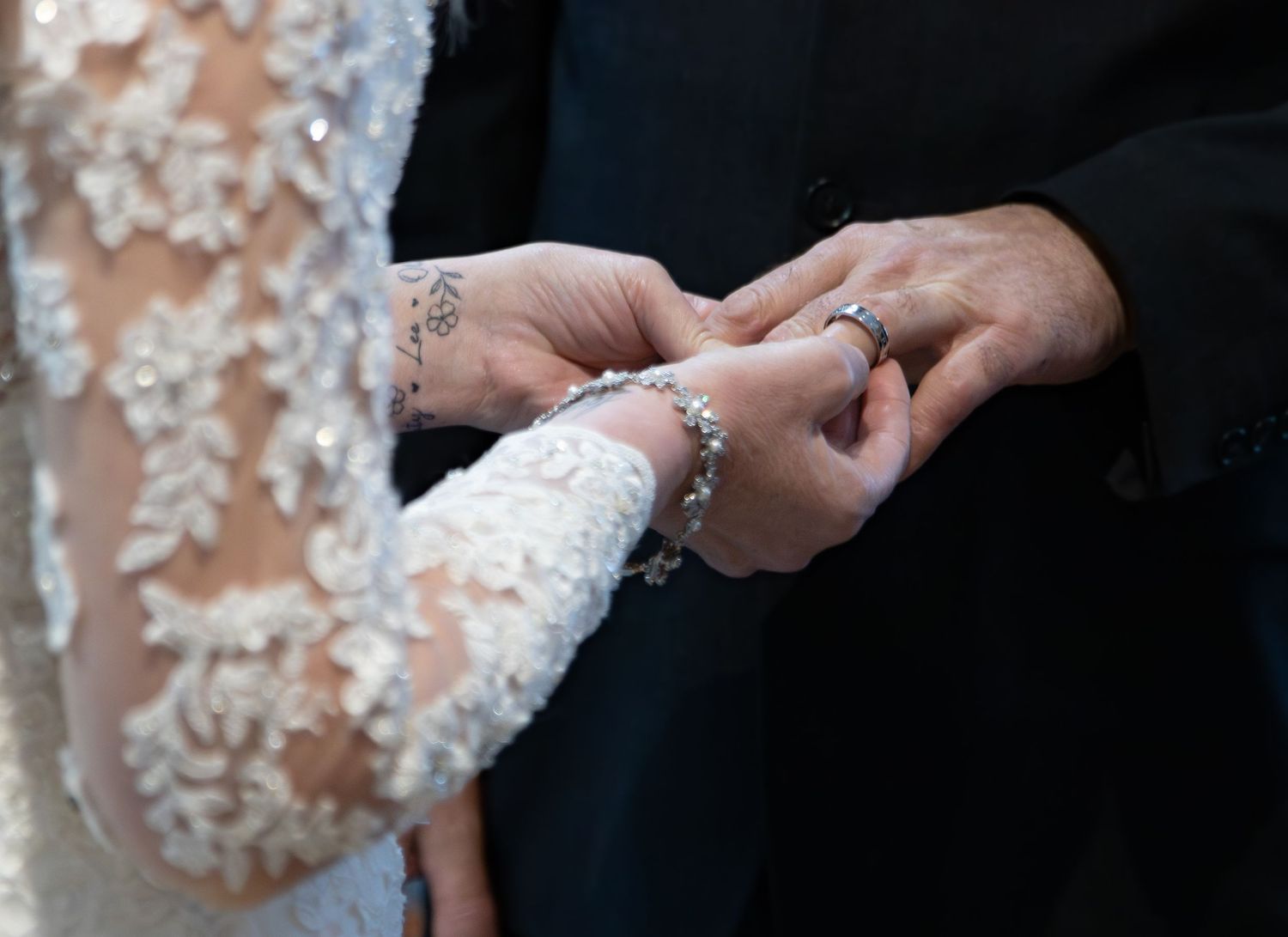 Hands exchanging wedding rings during ceremony with white lace sleeve detail visible.