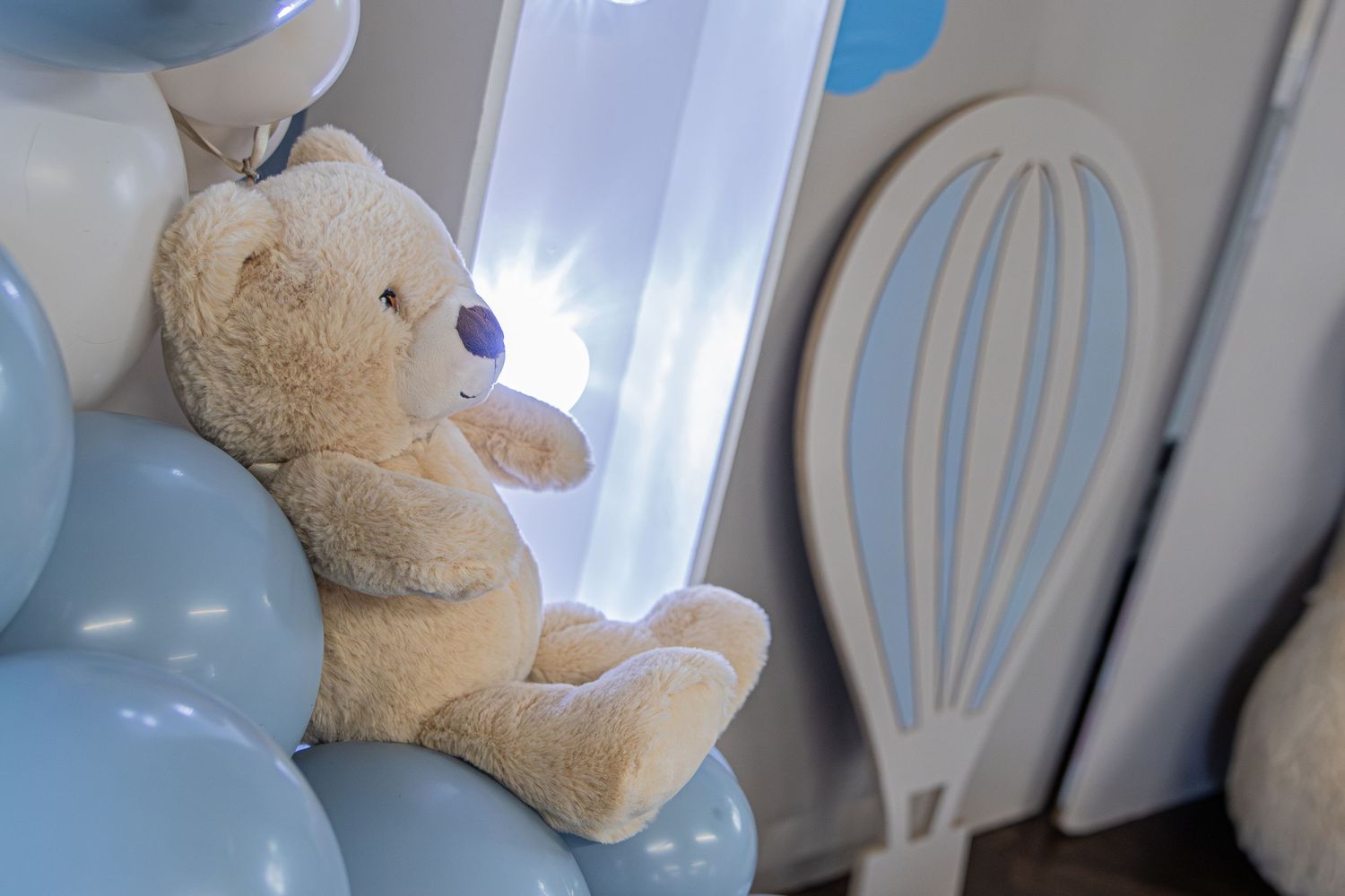 Teddy bear sitting among light blue balloons next to a decorative hot air balloon prop in a baby shower setup.