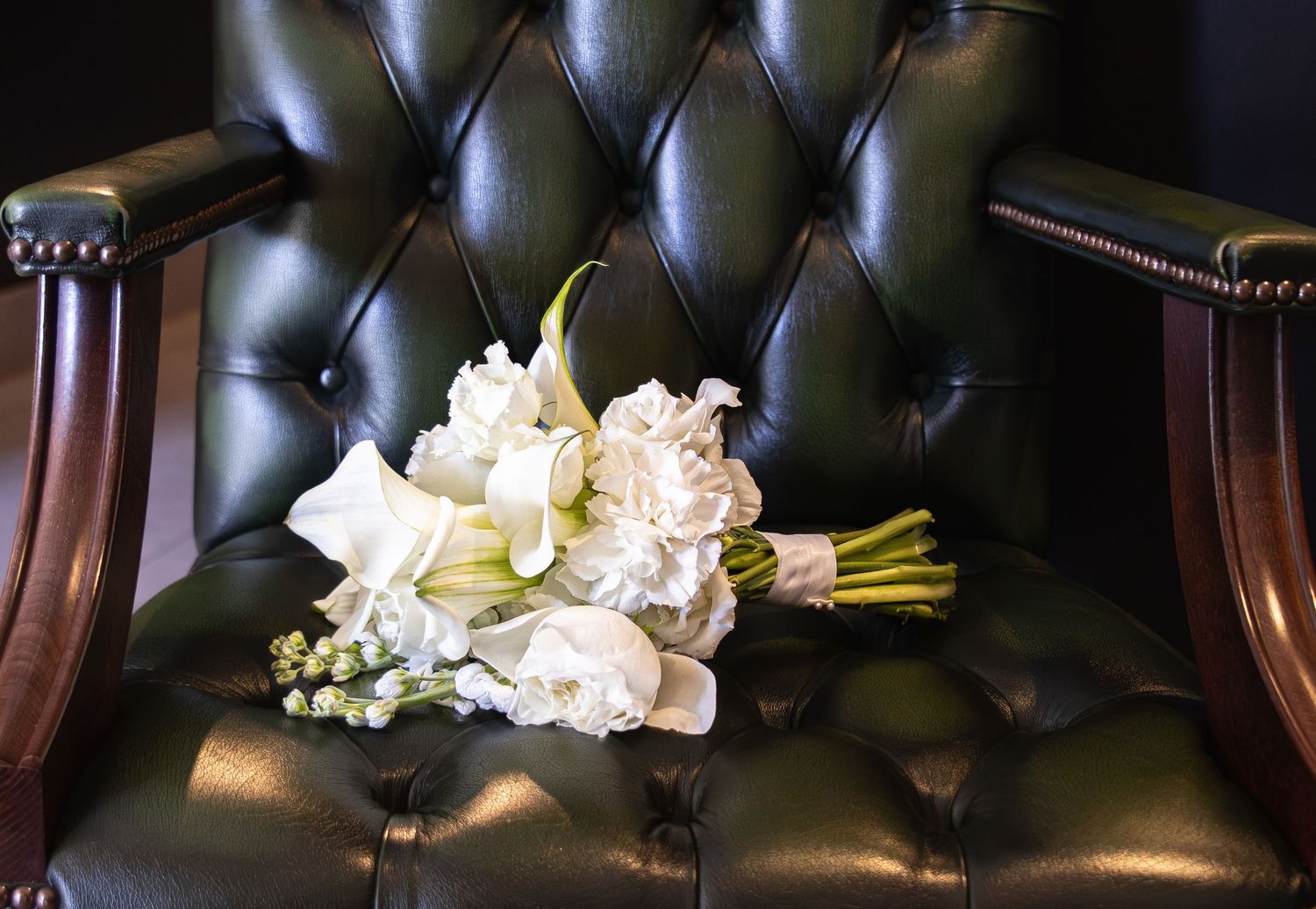 White bridal bouquet of roses and calla lilies resting on a dark green tufted leather chair.