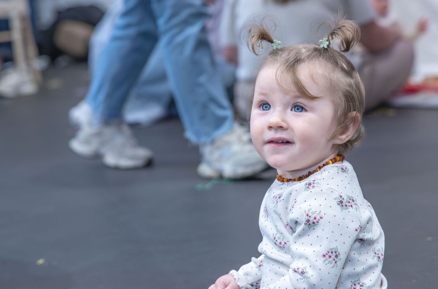 Young toddler with hair buns sitting on pavement looking up while adults stand in background.