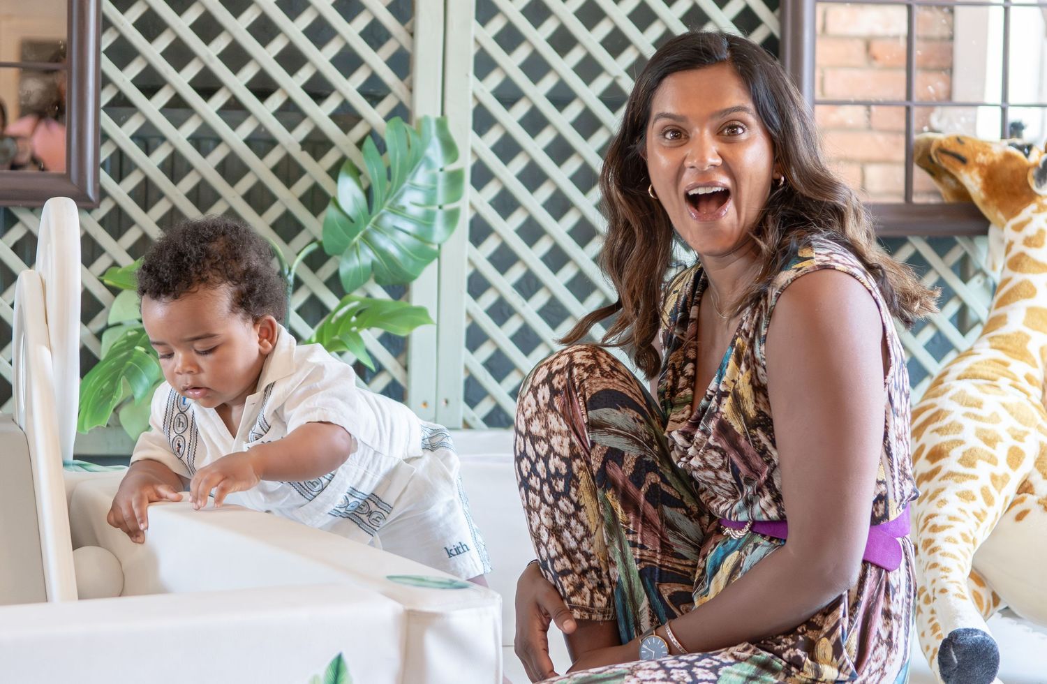 Woman in colorful dress laughing with toddler at white table, giraffe toy in background.