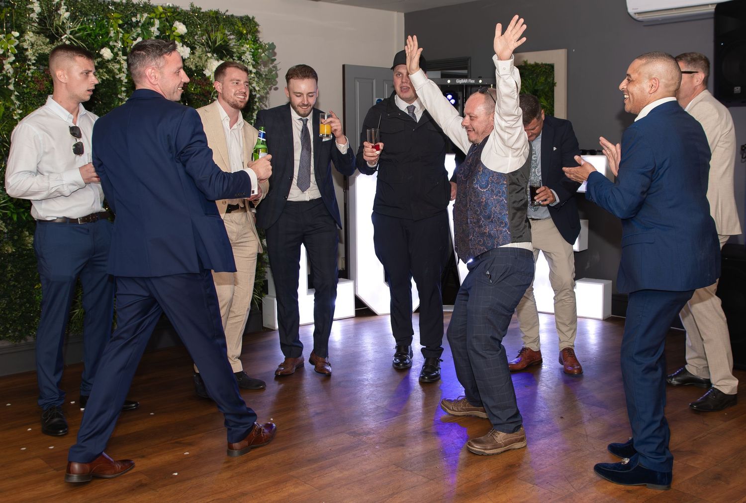 Men in suits dancing and celebrating at a formal event or wedding reception indoors.