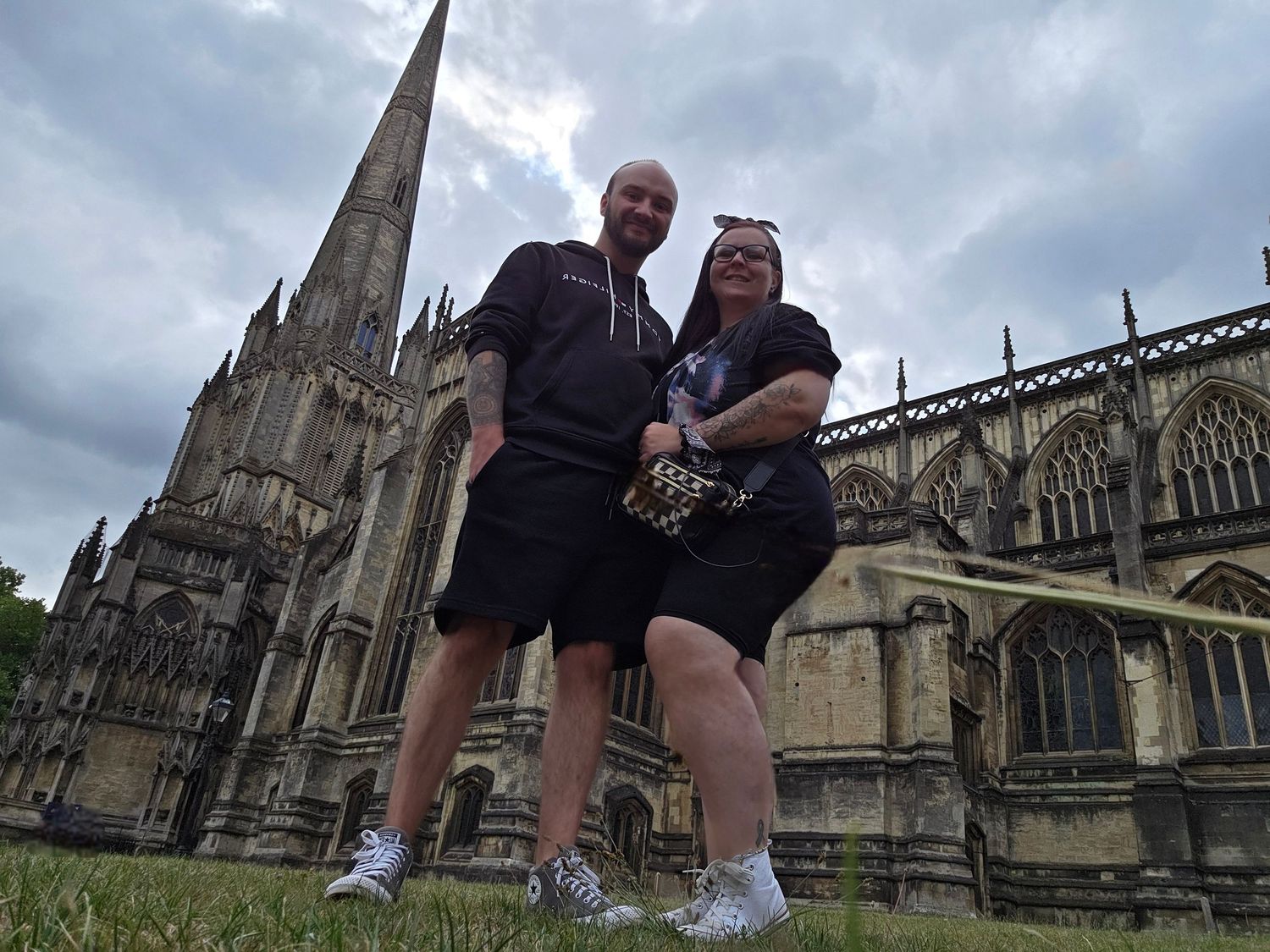 A couple poses outside a Gothic cathedral with dramatic spires and architecture under cloudy skies.