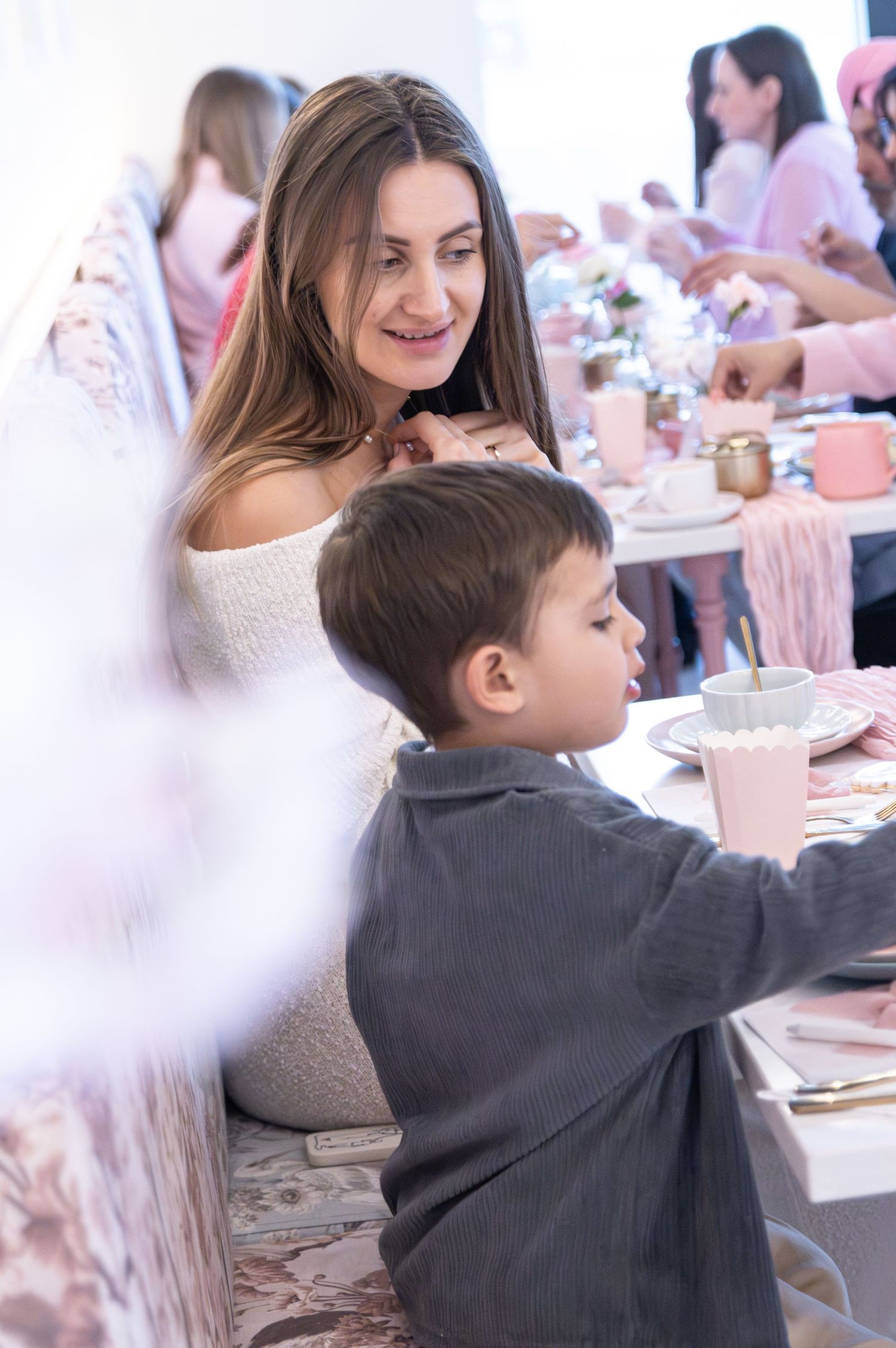A woman smiles warmly at a young boy during an elegant pink-themed party with decorated tables in the background.