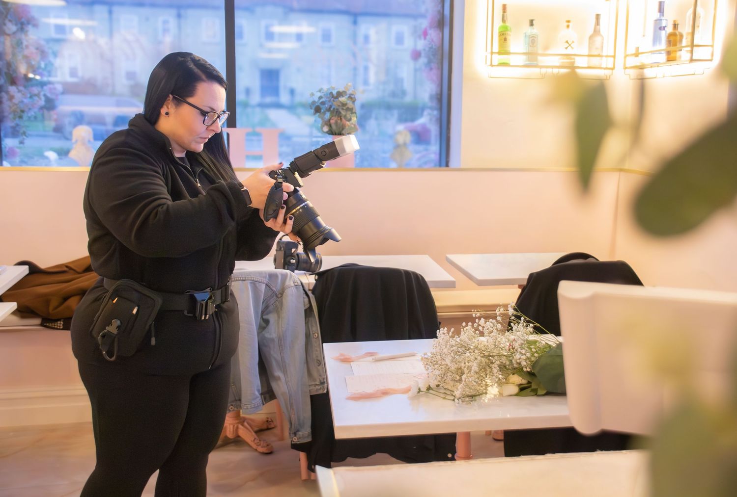 Photographer in black outfit adjusting camera lens indoors at a bright, modern studio space with floral decor.