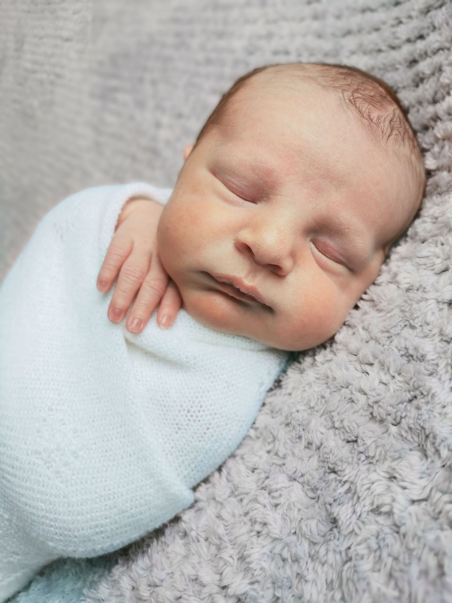 Peaceful sleeping newborn baby swaddled in a light blue wrap on a soft gray blanket.